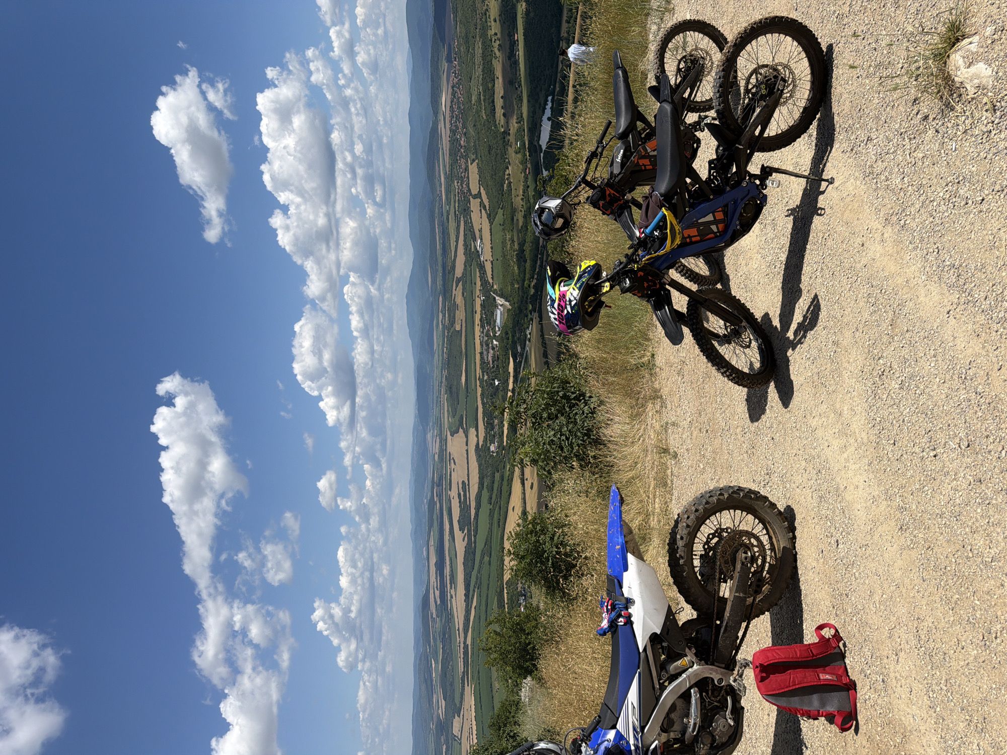 This image depicts a scenic overlook with two dirt bikes parked on a gravel path. The foreground features the rear section of a blue and white dirt bike, with a red backpack resting on the ground next to its rear wheel. To the right, another dirt bike, primarily black and blue, is parked with a helmet resting on its handlebars. In the background, a vast landscape unfolds, characterized by rolling hills, agricultural fields, and distant mountains under a partly cloudy blue sky. The location is identified as the city of Sheremetia, Bulgaria.