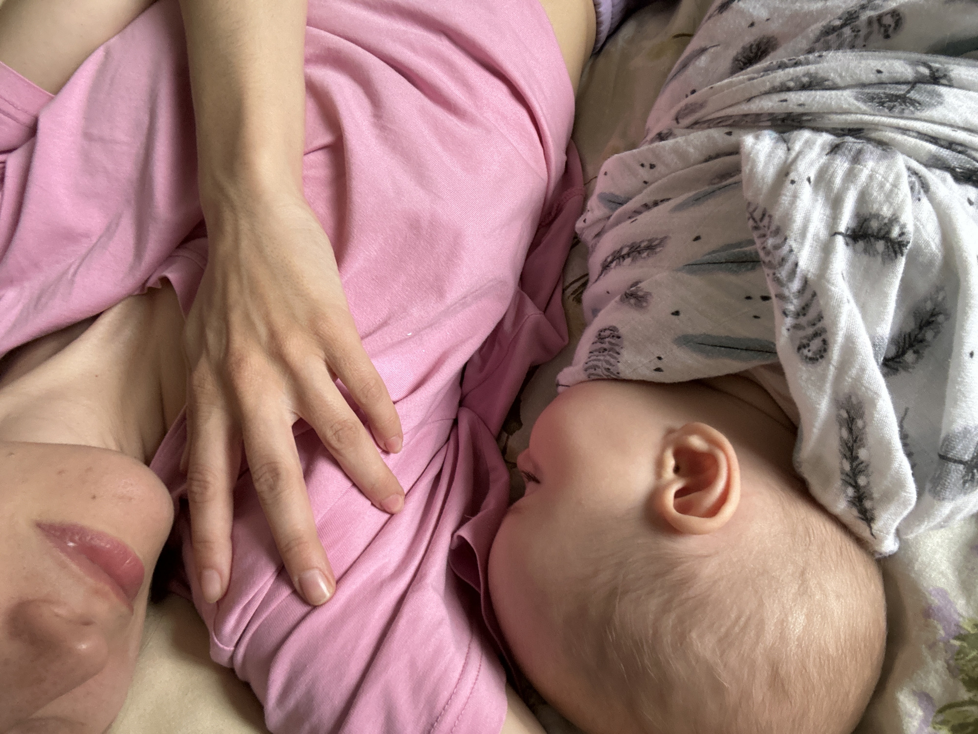 A top-down view depicts a person lying beside a baby. The person is partially visible from the side, revealing their face and hand resting on their pink-colored clothing. The baby lies close, covered in a patterned white blanket with tree-like designs. The baby's ear and head are visible. The background seems to be a bed with a patterned sheet. No location information about Moscow, Russia, can be discerned from the image.