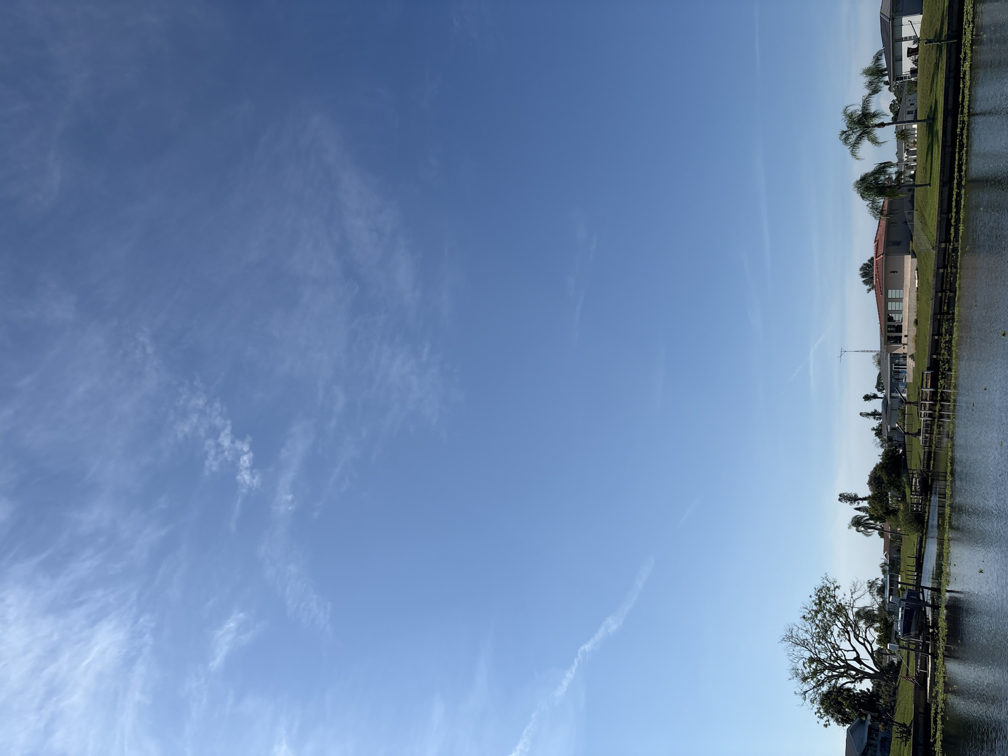 This media file depicts a serene canal-side view in North Port, USA, likely during the daytime under clear skies. The scene is dominated by a vast expanse of blue sky, dotted with wispy white clouds. On the right side of the frame, a tranquil body of water reflects the sky and the surrounding landscape.
The shoreline is lined with lush green grass and features several houses with tiled roofs, characteristic of a residential canal community. Palm trees and other varied foliage add to the tropical ambiance. A boat dock is visible, suggesting recreational activities. In the foreground, a large, mature tree with a sprawling canopy stands prominently. The overall atmosphere is peaceful and idyllic, evoking a sense of calm and relaxation. The clear sky indicates fair weather, and the presence of boats hints at a leisurely setting. No visible text is present in the media file.