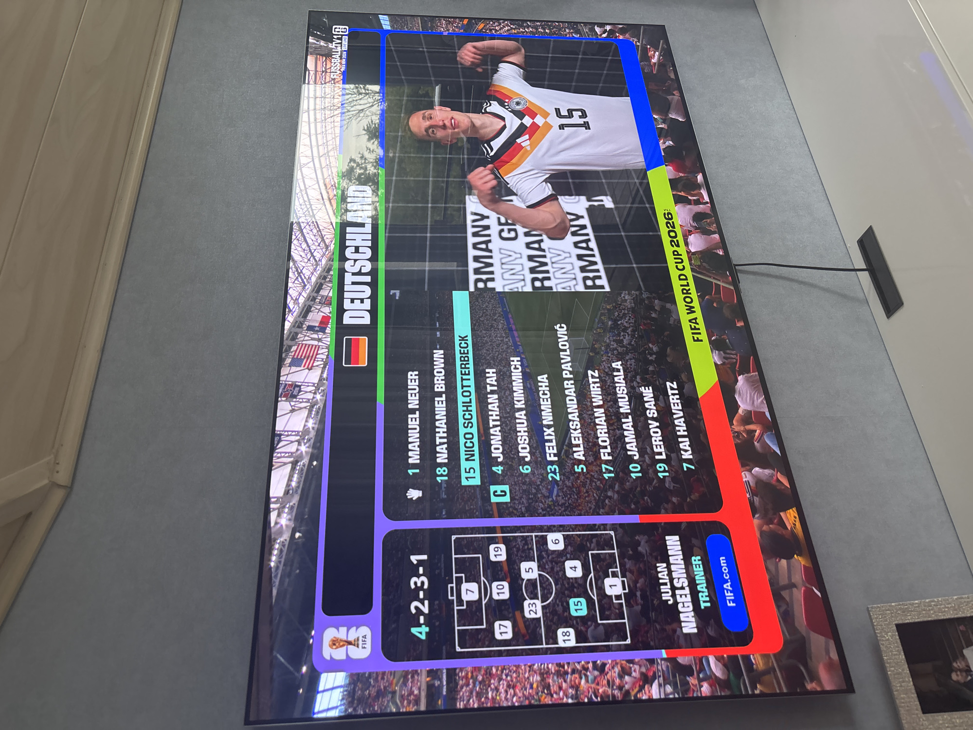 The image shows a television screen displaying a football match lineup for Germany. The screen shows the formation "4-2-3-1", the German flag, the word "DEUTSCHLAND", and a list of players with their jersey numbers. The players listed are Manuel Neuer (1), Nathaniel Brown (18), Nico Schlotterbeck (15), Jonathan Tah (4), Joshua Kimmich (6), Felix Nmecha (23), Aleksandar Pavlovic (5), Florian Wirtz (17), Jamal Musiala (10), Leroy Sané (19), and Kai Havertz (7). Julian Nagelsmann is listed as the trainer. A player in a German jersey with the number 15 on it is also shown on the right side of the screen. The bottom of the screen displays "FIFA WORLD CUP 2026". The television is placed on a white surface in front of a grey textured wall, with a silver picture frame visible on the left.