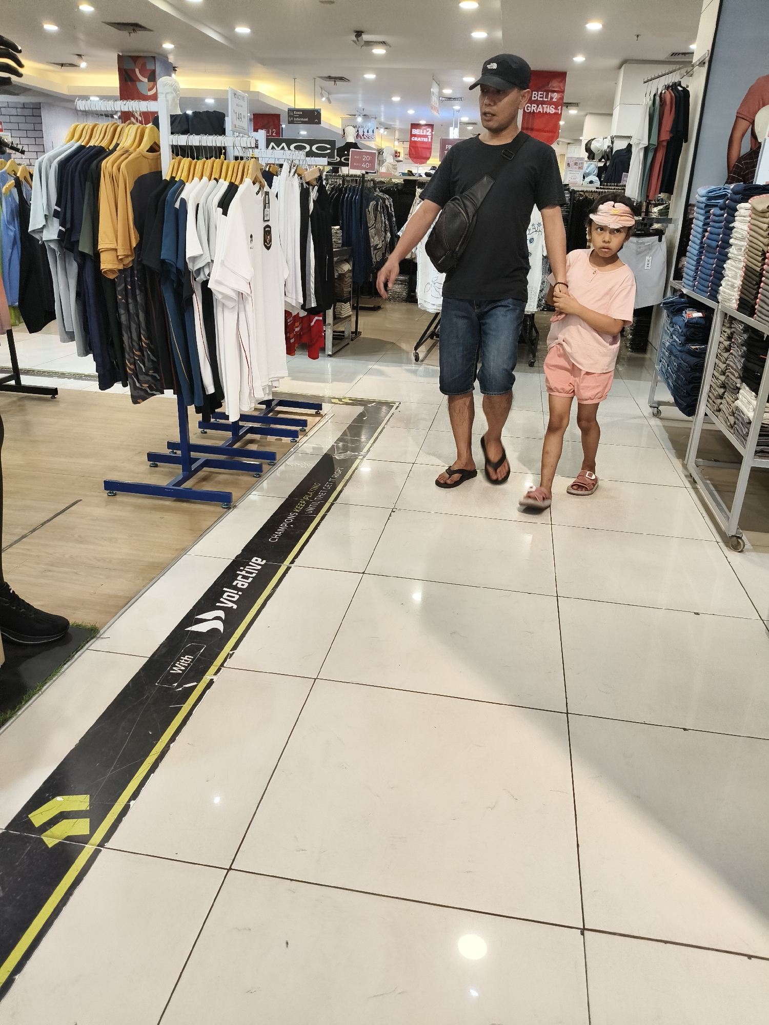 A man and a young girl are walking in a clothing store. The man is wearing a black t-shirt, blue denim shorts, and a black baseball cap. He has a black crossbody bag and is wearing black flip-flops. The girl is wearing a pink t-shirt, pink shorts, and pink sandals, and a pink cap. The man is holding the girl's hand. They are walking on a tiled floor with a black and yellow stripe on the floor that says "yo! active CHAMPIONS KEEP PLAYING UNTIL THEY GET IT RIGHT". In the background, there are racks of clothing and other store fixtures. The lighting is typical of a retail environment. The image was taken in the city of Bandung, Indonesia, likely within a department store such as Matahari or Ramayana, given the common retail signage and clothing displays found in such locations in Indonesia.