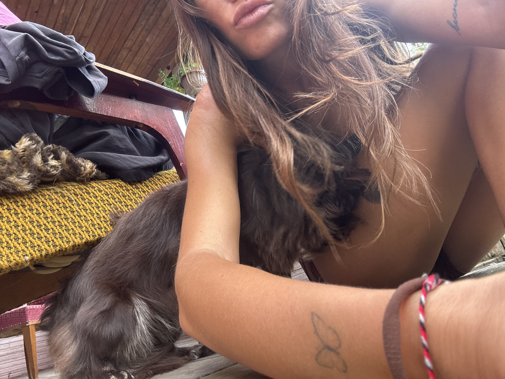 A woman is sitting on a wooden deck with a dog. The woman has long brown hair and is wearing a bikini top. The dog is dark brown with some graying fur. There is a wooden chair with clothes on it in the background. The scene is outdoors, likely on a porch or patio. The image was taken in Krasnaya Presnya SNT, Russia.