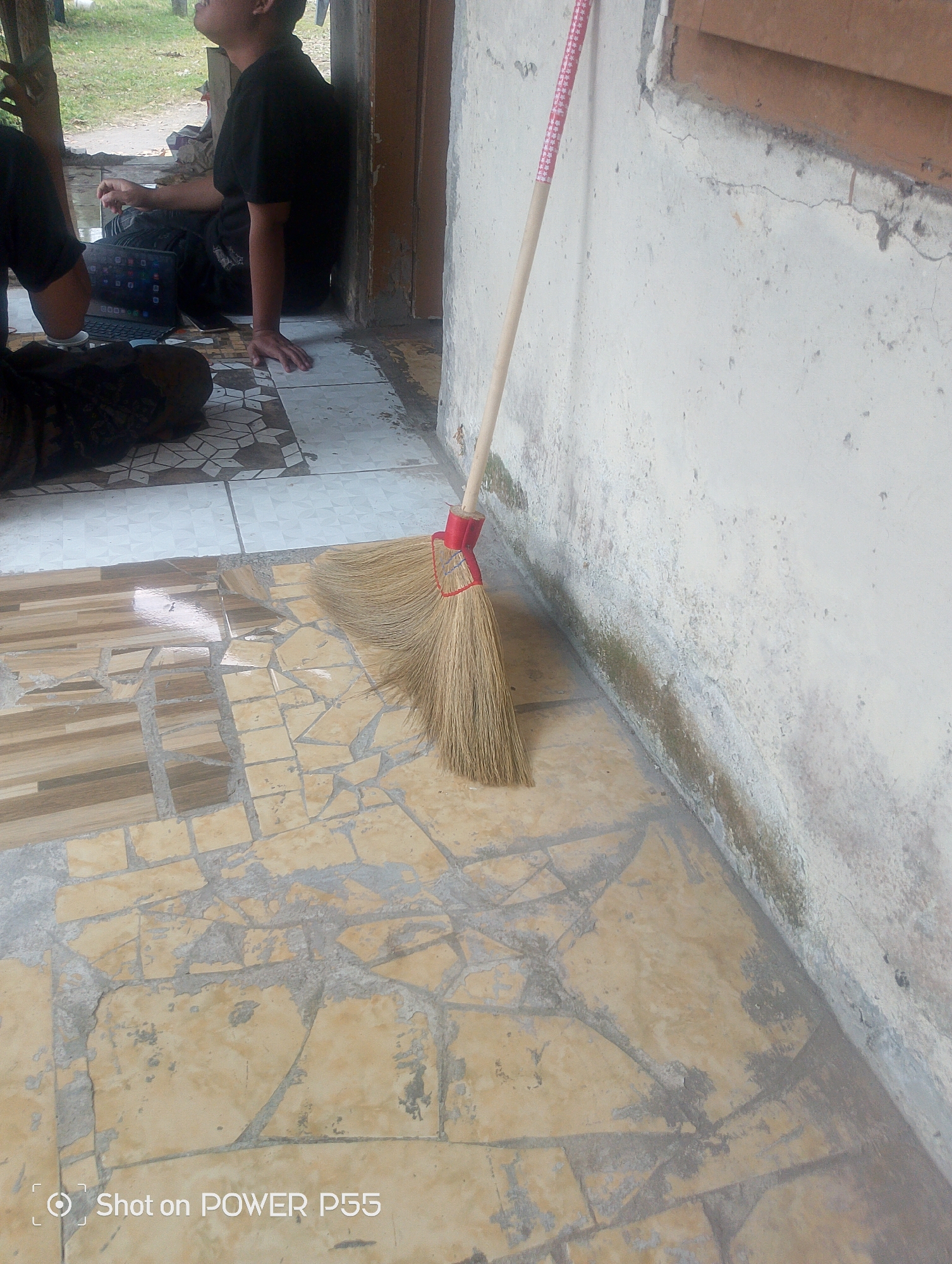 A close-up, low-angle shot focuses on a broom leaning against a weathered, off-white concrete wall. The broom's natural, straw-like bristles are fanned out on a textured tiled floor, which is a mosaic of tan and brown rectangular tiles interspersed with irregularly shaped, broken pieces of tile and visible grout. To the left of the broom, a section of a person wearing a black shirt is visible, along with a dark fabric object and a laptop with its screen illuminated. The background shows a glimpse of lush green grass and what appears to be a wooden structure. The image has a timestamp overlay in the bottom left corner: "Shot on POWER P55". The scene suggests an outdoor or semi-outdoor area, possibly a porch or a patio, with a candid and slightly worn aesthetic.