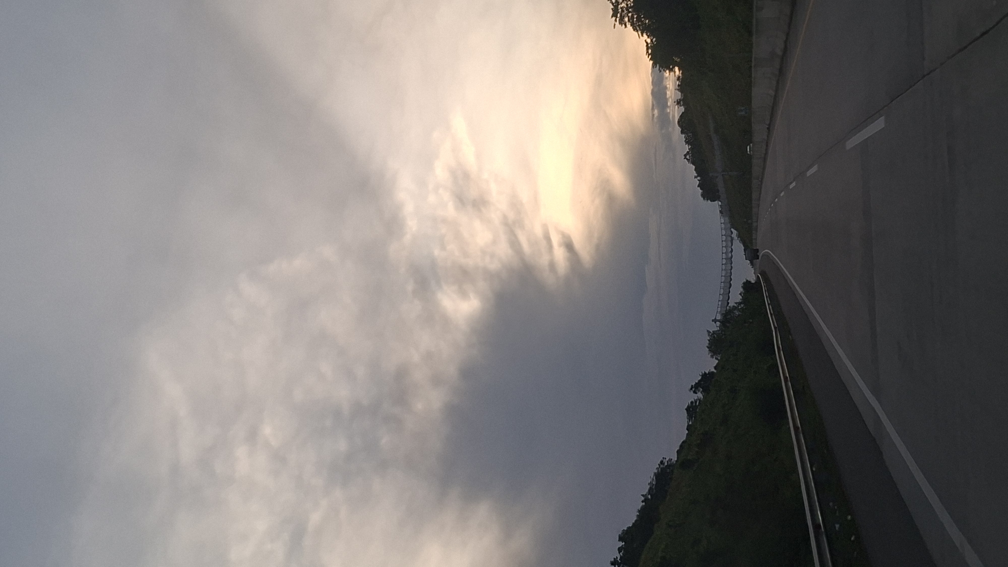 The image captures a highway in Lamtamot, Indonesia, under a dramatic sky. The road curves to the right, with a dark, forested hillside on the left and a lighter, less vegetated slope on the right. The sky is filled with heavy, gray clouds, but a bright, yellow-orange light breaks through on the right side, suggesting either sunrise or sunset. A suspension bridge spans across the highway in the distance, and a few vehicles are visible on the road. The overall impression is one of an overcast day on a scenic route.