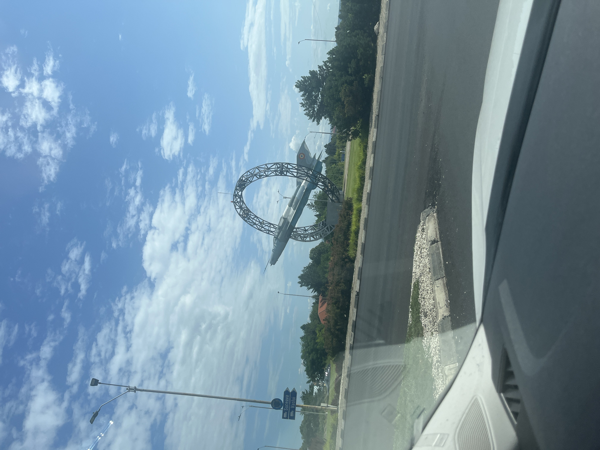 This media file shows an outdoor scene, likely in Bacău, Romania, on a clear, sunny day with scattered clouds. The primary object of focus is an airplane sculpture that appears to be suspended within a large, metallic loop structure. The airplane is painted in a grey and blue camouflage pattern with national insignia visible on its tail. This structure is situated near a road, with a white car visible in the foreground, suggesting the image was taken from inside a vehicle.
Behind the sculpture, a dense line of green trees forms a backdrop, with some red-tiled rooftops peeking through in the distance, hinting at a developed area. In the lower part of the image, there are streetlights and road signs. One sign with blue background and white text points to "SUCEAVA" and "VASLUI" with truck icons, indicating directions and possibly highway destinations. The road surface is asphalt, with a gravelly shoulder and a concrete curb.
The lighting suggests it is daytime, likely late morning or early afternoon, given the bright sunlight and clear sky. The overall impression is one of a roadside monument or landmark, possibly commemorating aviation history or serving as a local attraction. There are no people visible in the image. The scene conveys a sense of open space and a bright, pleasant atmosphere.