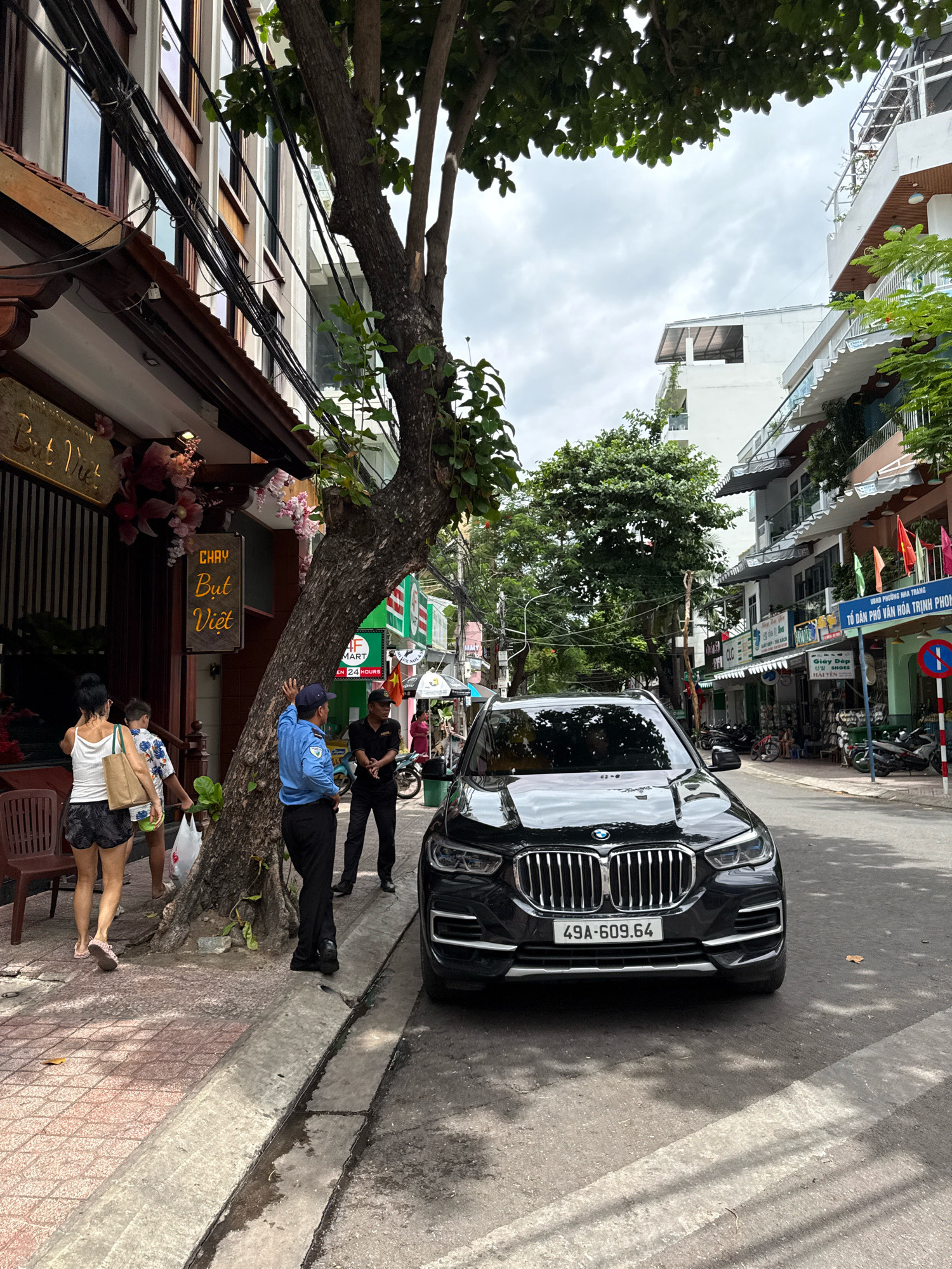 In Nha Trang, Vietnam, on a street lined with multi-story buildings, a black BMW SUV is parked on the right side of the road. A security guard in a blue uniform stands near the car, facing left and gesturing with his right hand. A man in a black shirt and pants, also appearing to be a guard, stands behind him, facing forward. Two women and a child are walking on the sidewalk to the left of the parked car, away from the camera. A large tree with a thick trunk is positioned between the sidewalk and the street, partially obscuring the view of the buildings. The street appears to be a commercial area with shops and signs visible in the background. The sky is overcast.
