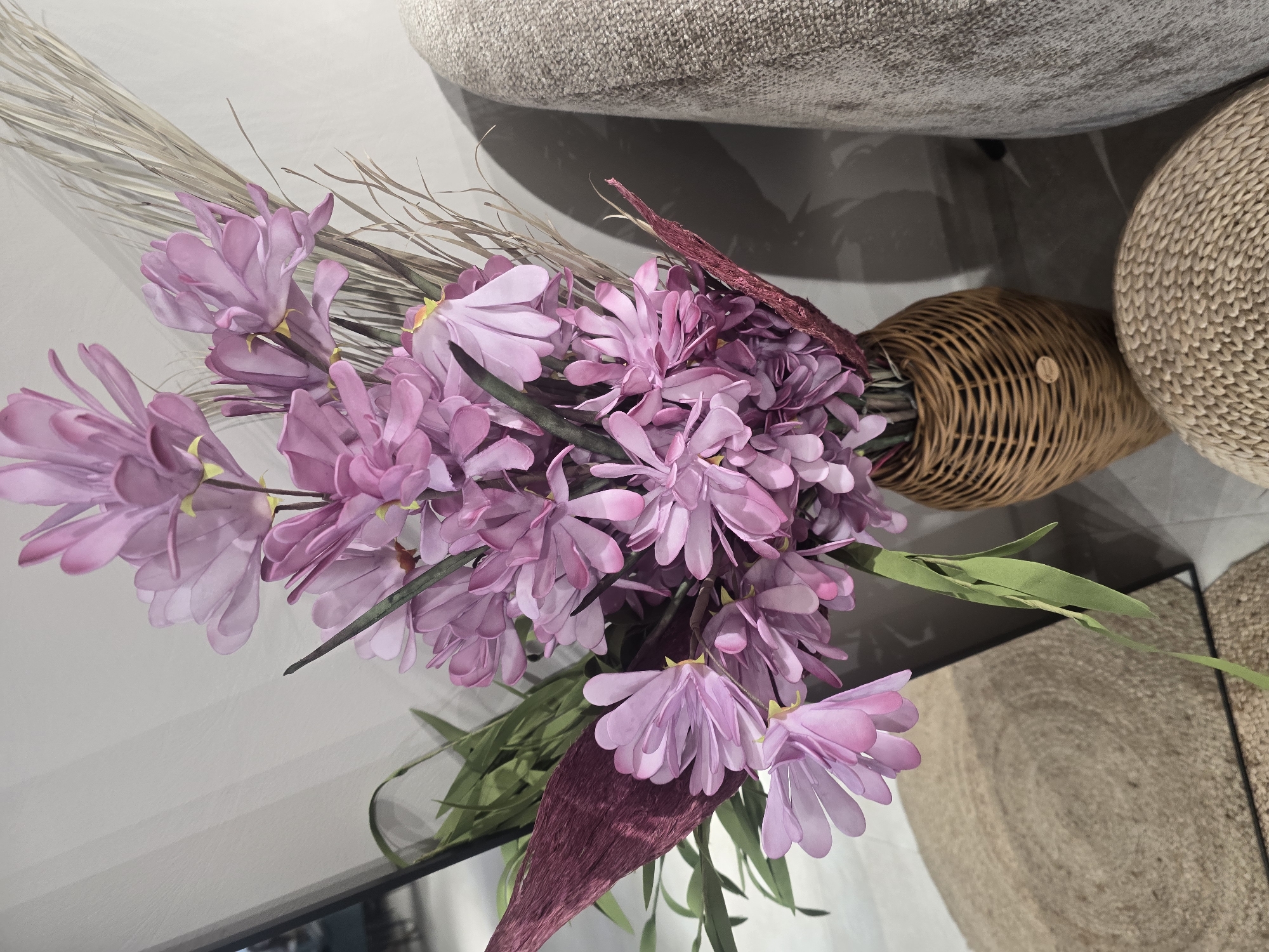 A close-up, eye-level shot focuses on a woven vase filled with a bouquet of artificial pink flowers and dried grass. The vase is made of woven natural fibers and has a rounded shape, tapering towards the top. The flowers are delicate with multiple petals and have yellow accents in the center. Among the flowers are thin, straw-like dried grasses, adding texture to the arrangement. Some deep red, feathery decorative elements are also interspersed within the bouquet. The background shows a light-colored, vertically striped wall, and to the right, a portion of a textured, neutral-colored upholstered piece of furniture is visible. In the lower left of the frame, a circular, woven mat made of natural fibers is partially visible. The scene appears to be an indoor setting, possibly a home or commercial space. There is no indication of the specific location within Ulan-Ude, Russia.