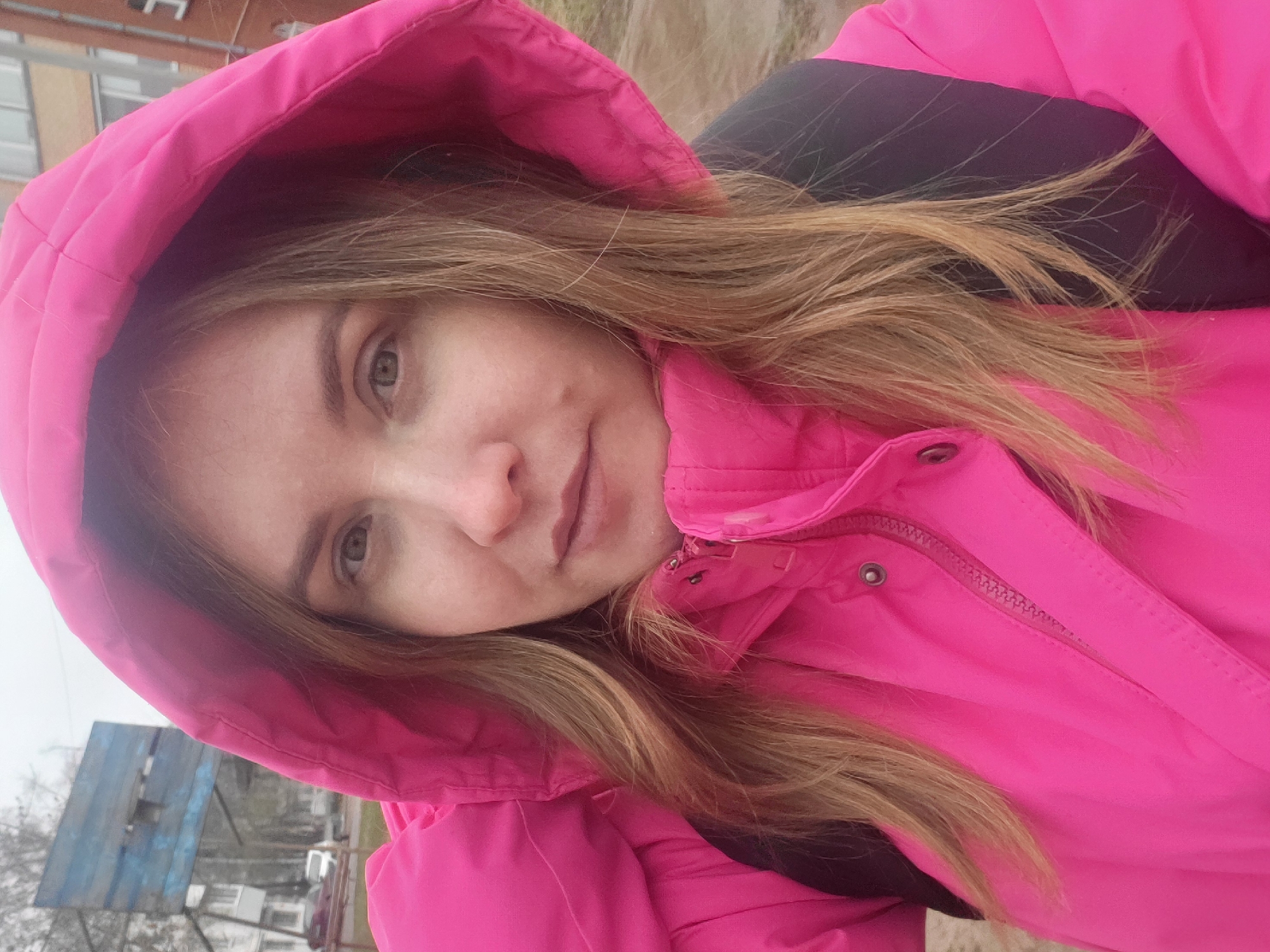 A medium close-up, slightly tilted selfie captures a young woman outdoors. She is wearing a bright fuchsia pink hooded jacket, with the hood up around her head. Her long, light brown hair falls around her face and over her shoulders, some strands visible against the black interior lining of her jacket. She has light-colored eyes (likely green or hazel) and a calm, slightly smiling expression. Her gaze is directed towards the camera.
The background is somewhat blurred but reveals an overcast sky and muted colors. On the left side, a weathered, faded blue structure resembling a basketball backboard is visible. Behind it, glimpses of buildings and possibly trees suggest an urban or suburban setting. The overall lighting is soft and diffused, indicating an overcast day. No specific time of day is clearly discernible, but it appears to be daylight. There is no visible text in the image. The scene suggests a casual outdoor moment in Chaikovsky, Russia.