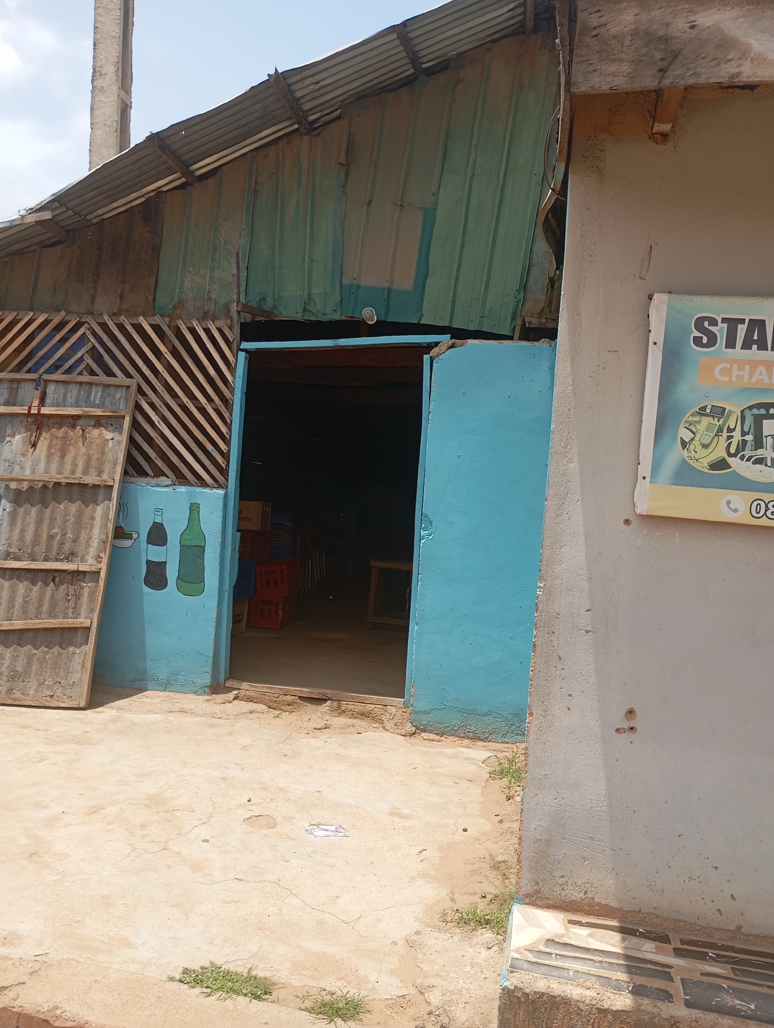 A weathered, corrugated metal roof supported by wooden beams covers a structure with light blue painted walls and a dirt ground. The entrance to the building is an open doorway with bright blue double doors. To the left of the entrance, a faded mural depicts two bottles and a bowl of food painted on the blue wall. A leaning, rectangular wooden structure with corrugated metal panels stands to the left of the mural. A sign is visible on the right wall, featuring the text "STAR" and "CHAI" along with circular graphics depicting various items. The overall scene is that of a small, possibly informal, commercial establishment in Shendam, Nigeria.