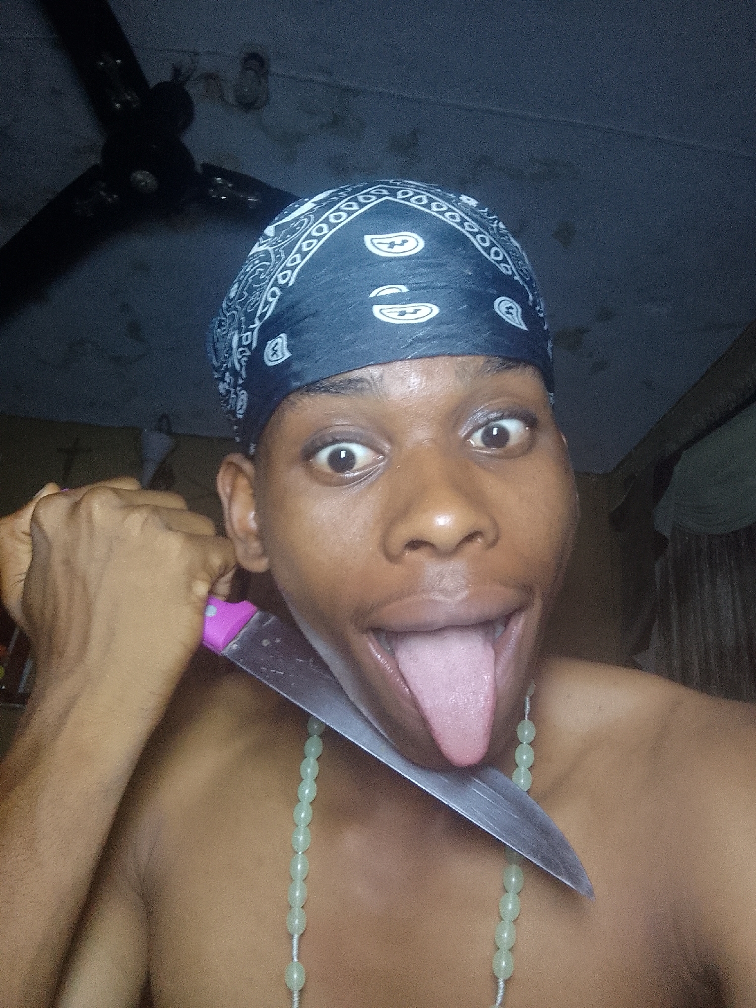 A dark-skinned individual wearing a blue paisley bandana and a string of light green beads around their neck is holding a knife to their throat, with their tongue sticking out. The location is unknown. In the background, there is a ceiling fan.