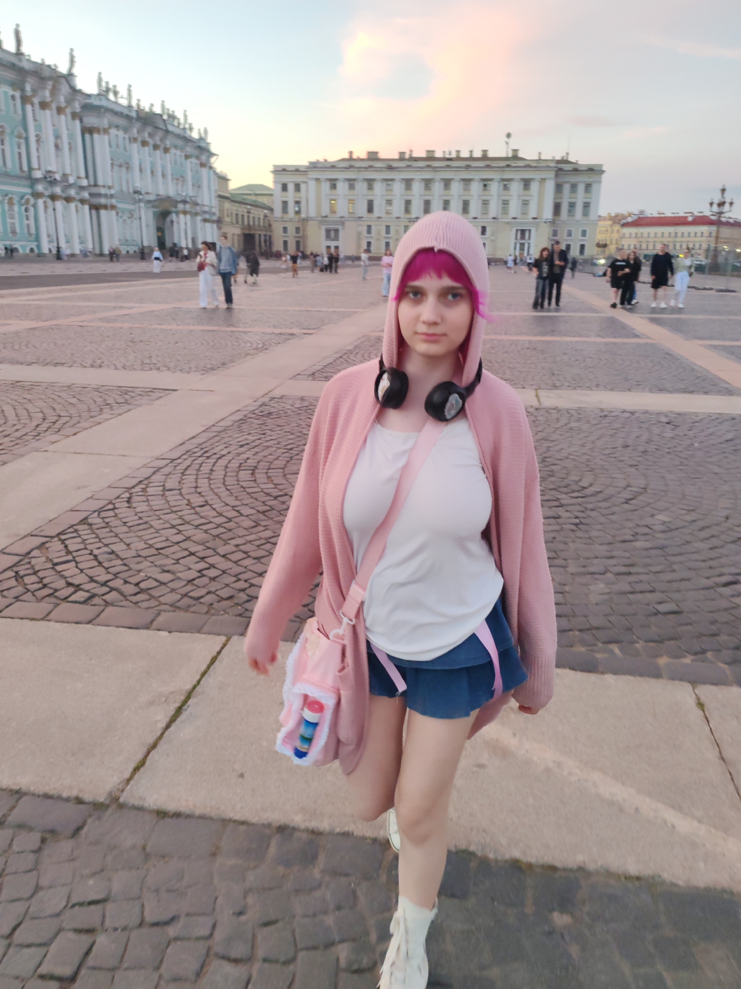 In this full-body, eye-level shot, a young person with vibrant pink hair and headphones around their neck walks towards the viewer in Palace Square, Saint Petersburg, Russia. The individual wears a light pink hooded cardigan over a white t-shirt and a denim skirt, with white sneakers and socks. A pink shoulder bag with white trim hangs on their right side, containing a blue and red object. The square's cobblestone and paved ground is visible, with several other people walking in the background. Behind them, the grand facades of the Winter Palace and the General Staff Building create an imposing backdrop under a pale sky with hints of pink and yellow clouds, suggesting either dawn or dusk.