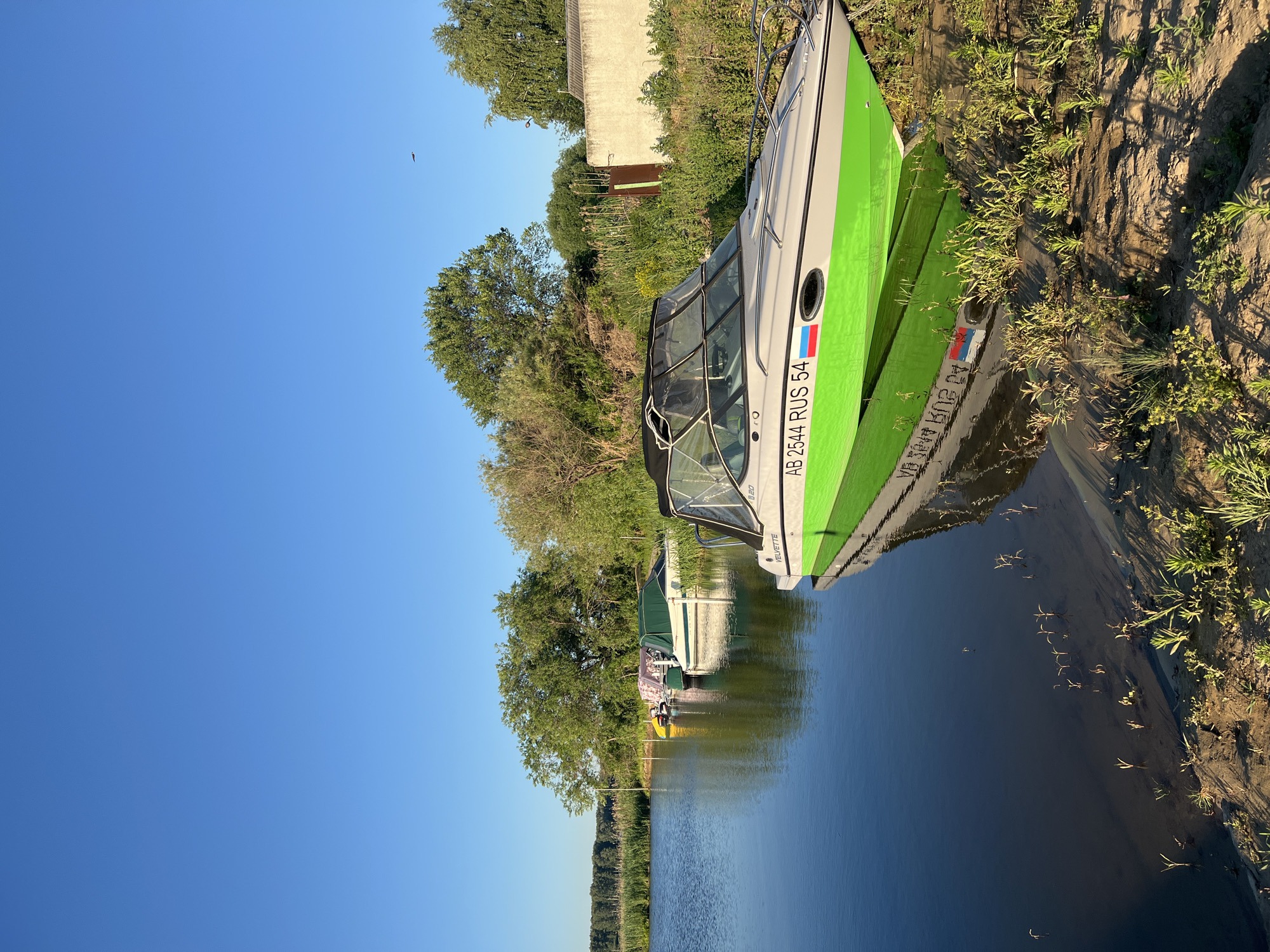 The image shows a bright, sunny day at a marina in Novopichugovo, Russia. A light-colored motorboat with a bright green hull is moored at the edge of a calm, dark body of water. The boat has a black canopy and a license plate that reads "AB 2544 RUS 54". The boat is reflected in the water, as are several other boats moored further down the shoreline. The background consists of lush green trees and vegetation, with a light-colored building visible on the right. The sky is a clear, vibrant blue, with a small bird visible in flight. The shoreline is sandy and covered with sparse green vegetation.