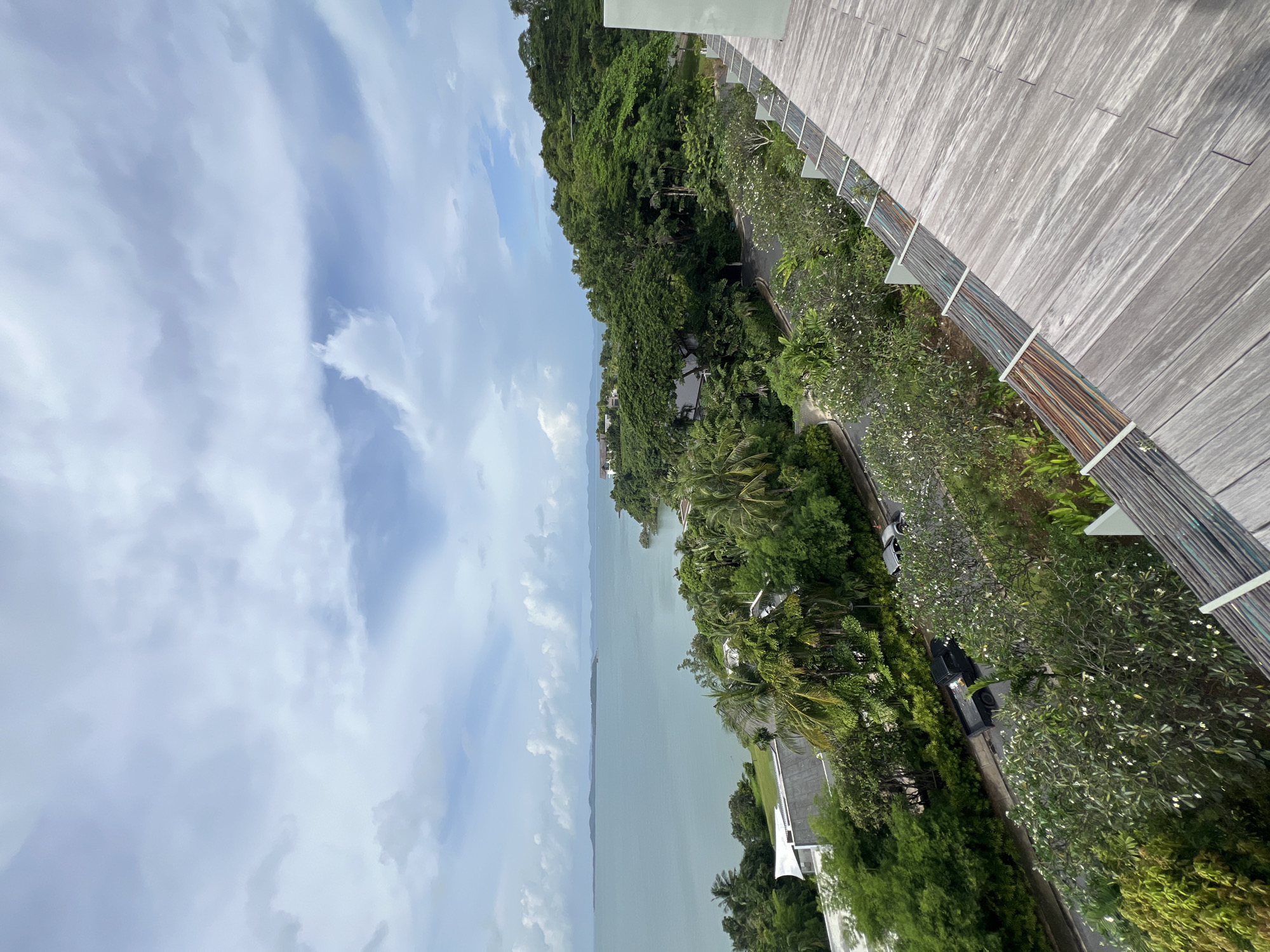 The image is a high-angle, wide shot of a coastal landscape in Pa Khlok, Thailand. In the foreground, a weathered wooden deck with visible wiring runs along the right side of the frame. Beyond the deck, lush green trees and tropical foliage dominate the scene, with some buildings partially visible amidst the vegetation. A paved road winds through the greenery, with a black pickup truck and a silver car parked on its side. In the mid-ground, the turquoise ocean stretches out, meeting a distant, hazy coastline under a partly cloudy sky. The overall impression is one of a serene, tropical environment.