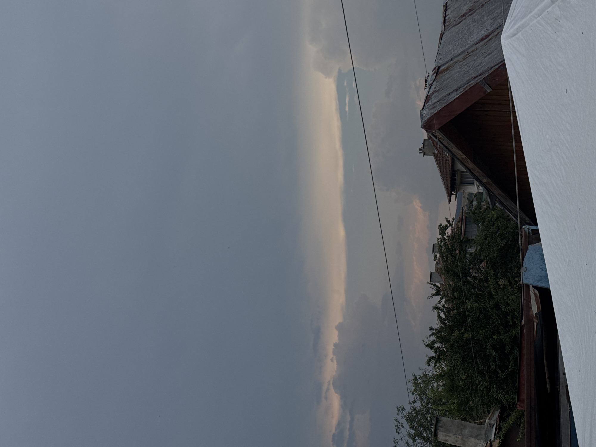 The image shows a cloudy sky over a residential area, likely in Sredets, Bulgaria, as indicated by the presence of houses with tiled roofs and chimneys, and dense trees. In the foreground, a white tarp covers some objects. A rusty metal roof of a house is visible, with wooden beams supporting it. The sky is overcast with dark gray clouds, but some lighter, pinkish hues suggest either sunset or sunrise. Electrical wires are strung across the scene. No people are visible.