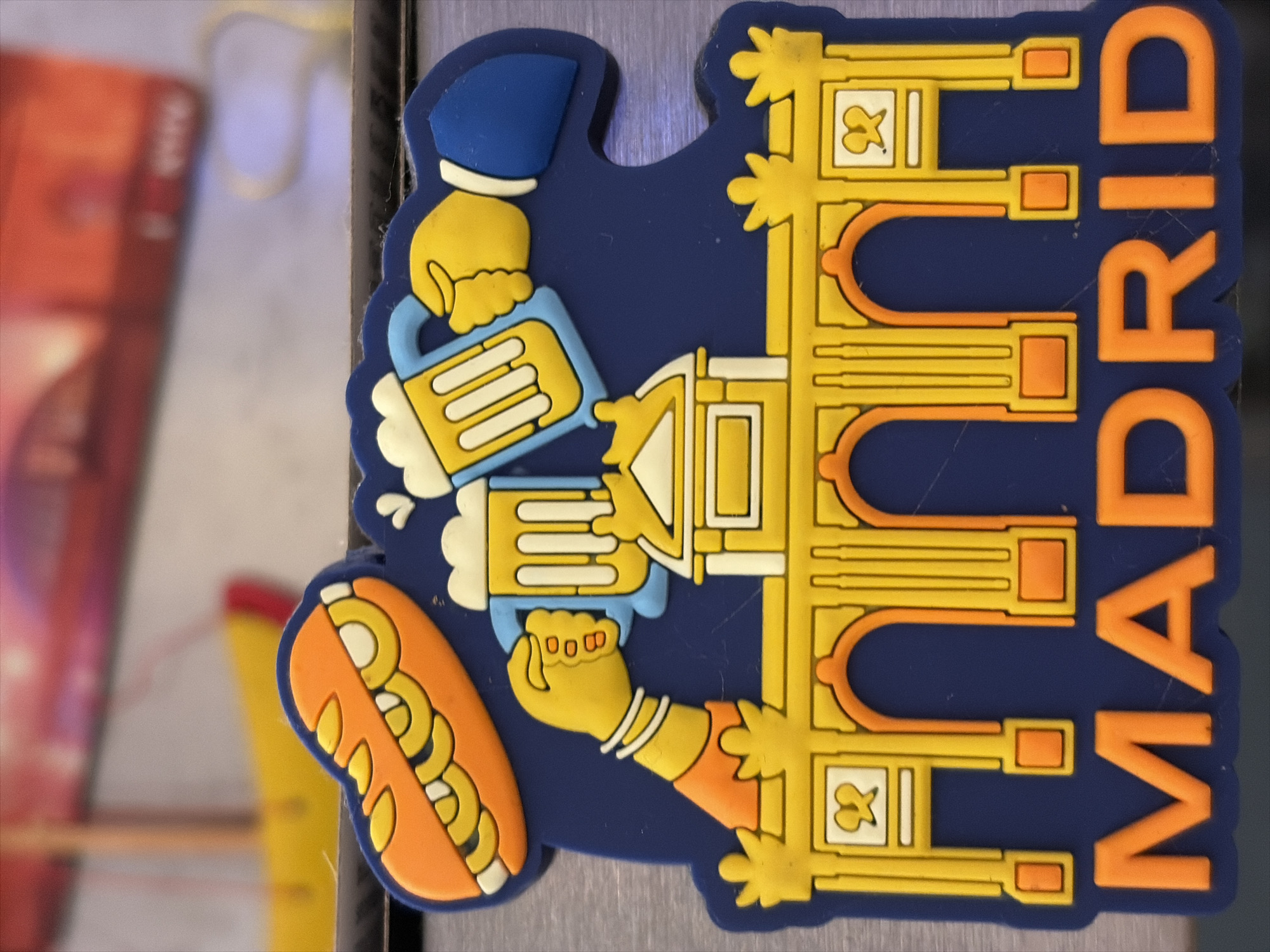 The image shows a dark blue magnet with a yellow and orange depiction of Madrid. At the top, an orange sandwich with yellow fillings is featured. Below the sandwich, two hands are shown raising mugs of beer. In the center, a yellow and white building resembling an archway is depicted. The word "MADRID" is written in large orange letters at the bottom of the magnet. The background is blurred and shows a hint of red and yellow objects, possibly part of a display or another magnet. There is no visible indication of Limnia, Northern Cyprus or Cyprus in the image.