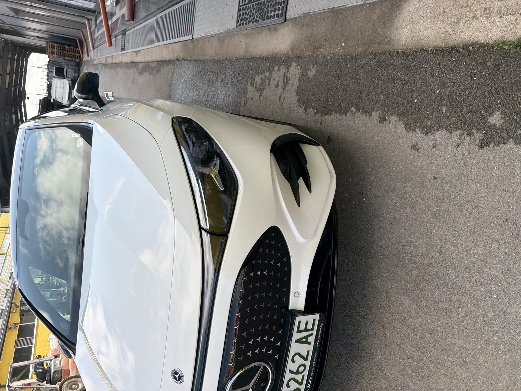 A white Mercedes-Benz car is prominently featured in the image, angled towards the viewer, with its front right section and part of the side and roof visible. The car appears to be stationary, positioned on an asphalt surface next to a concrete barrier or curb. The ground shows patches of wetness and varying textures of asphalt and concrete.
In the background, behind the car, various structures can be seen, suggesting an industrial area, a workshop, or a car service bay. These include metal grates on the ground, overhead structures, and in reflections on the car's surface, what appears to be a white van or truck and part of a yellow construction vehicle or lift. The sky, reflected in the car's windows and paintwork, appears overcast.
Notable details of the car include its modern design, the distinctive Mercedes-Benz logo on the grille and reflected on the hood, and intricate headlights. The grille features a unique pattern resembling many small three-pointed stars. A Bulgarian license plate is attached to the front bumper, displaying the characters "8262 AE." Below the license plate, text on the holder reads "Euro Stars - dealer and service" and "www.eurostars.bg," indicating a dealership or service center. The overall lighting suggests it is daytime. The presence of water marks on the ground could imply recent rain or the car being in a wash bay. The location context of Dolno Ablanovo, Bulgaria, aligns with the Bulgarian license plate and dealership information.
