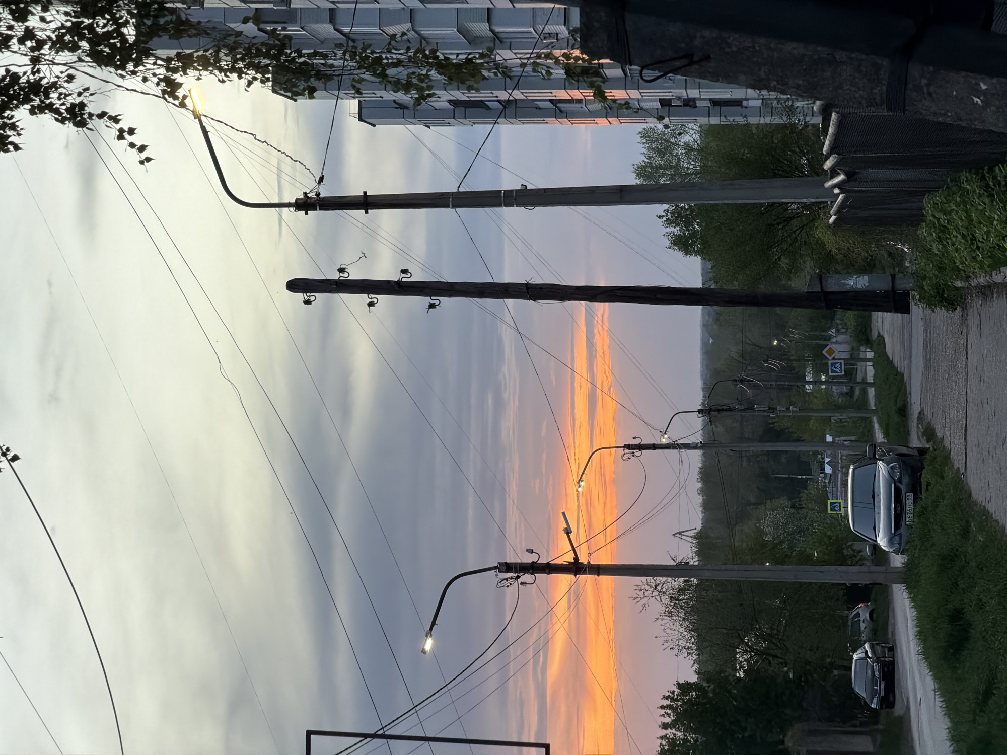 The image depicts a street in the city of Chaikovsky, Russia, at dusk. A line of utility poles runs alongside the road, with power lines crisscrossing the sky. Streetlights are attached to some of the poles, casting a glow. A gray car is parked on the right side of the road. The sunset casts a vibrant orange and purple glow on the sky. A building is visible on the left side of the road. Green vegetation is visible on both sides of the road. Road signs are seen in the distance.