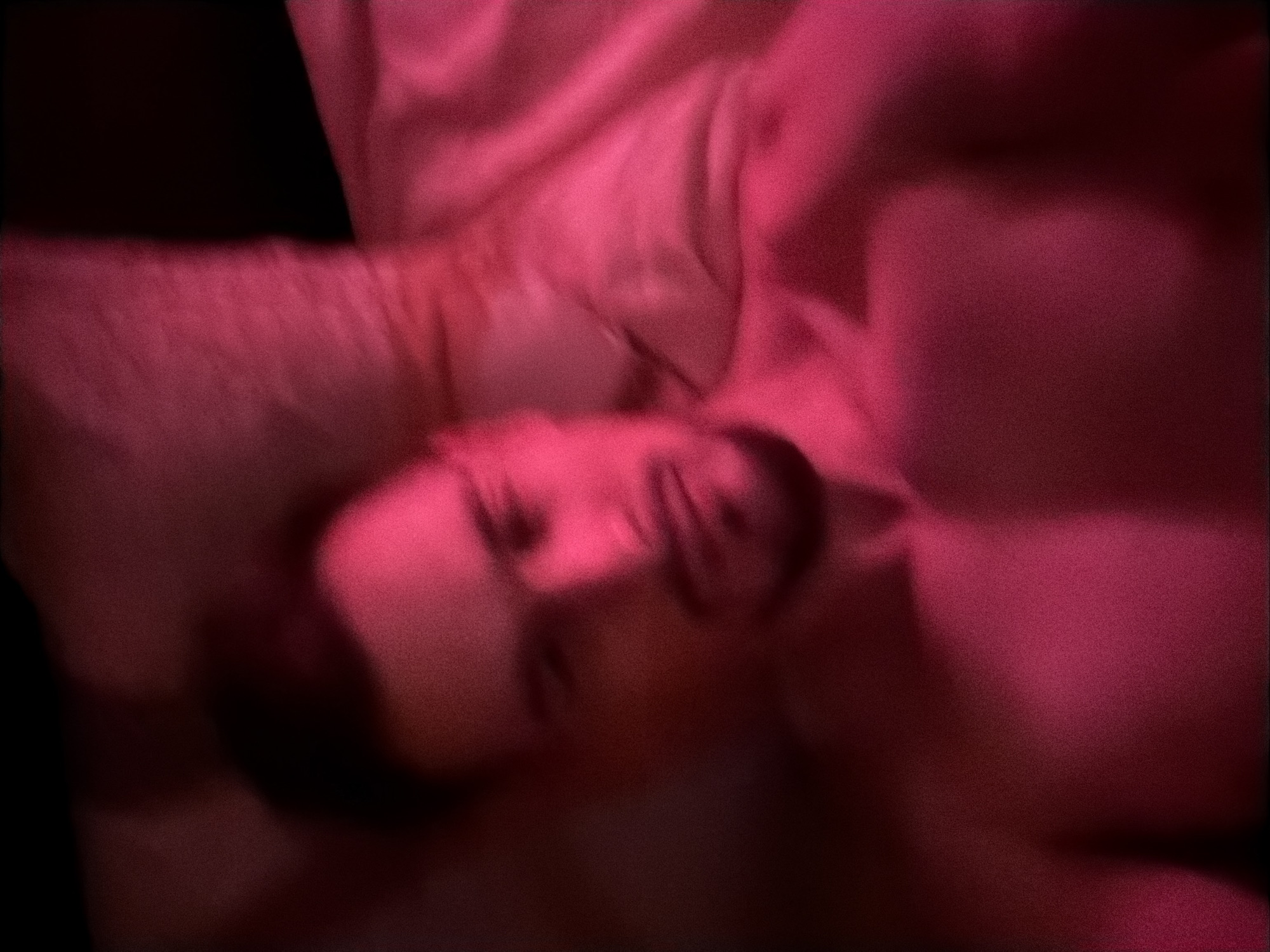 The image shows a blurred close-up of a person's upper body and face, illuminated by pink light. The person appears to be lying down, with their head resting on a pillow. Their chest and shoulders are visible. The background is mostly out of focus and also appears to be a pillow or bedding. There is no indication of the location in the city of Krasnoyarsk, Russia.
