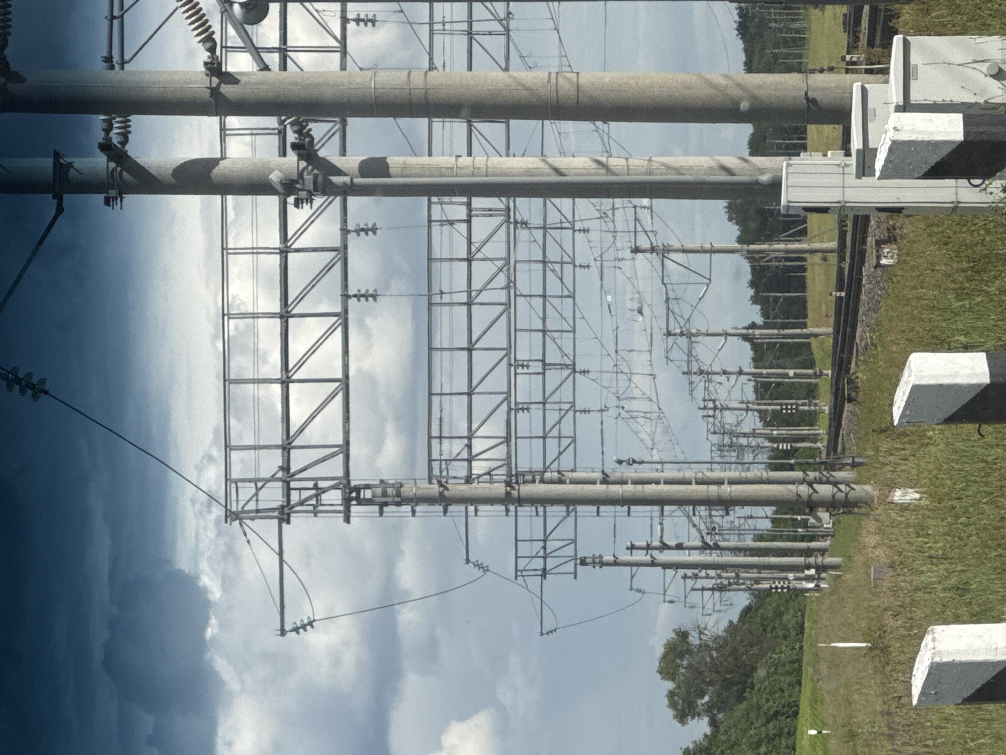 This is a landscape photograph taken outdoors, with the camera rotated 90 degrees counter-clockwise, so the image appears sideways. The primary subjects are a complex array of electrical transmission towers and equipment, characteristic of a power substation. Large, cylindrical grey conduits, likely high-voltage power lines, dominate the foreground, supported by tall metal lattice towers. Numerous smaller wires and insulators are visible, strung between the structures, creating a dense network against a cloudy sky. In the background, to the right, a grassy field stretches towards a treeline, with several concrete or stone markers, some with black and white stripes, placed along what appears to be a path or boundary. The weather appears overcast with dramatic, dark clouds on the left side of the image, contrasting with brighter, scattered clouds on the right. The overall impression is of a functional, industrial landscape under a dynamic sky, possibly a storm is approaching or passing. There is no visible text in the image.