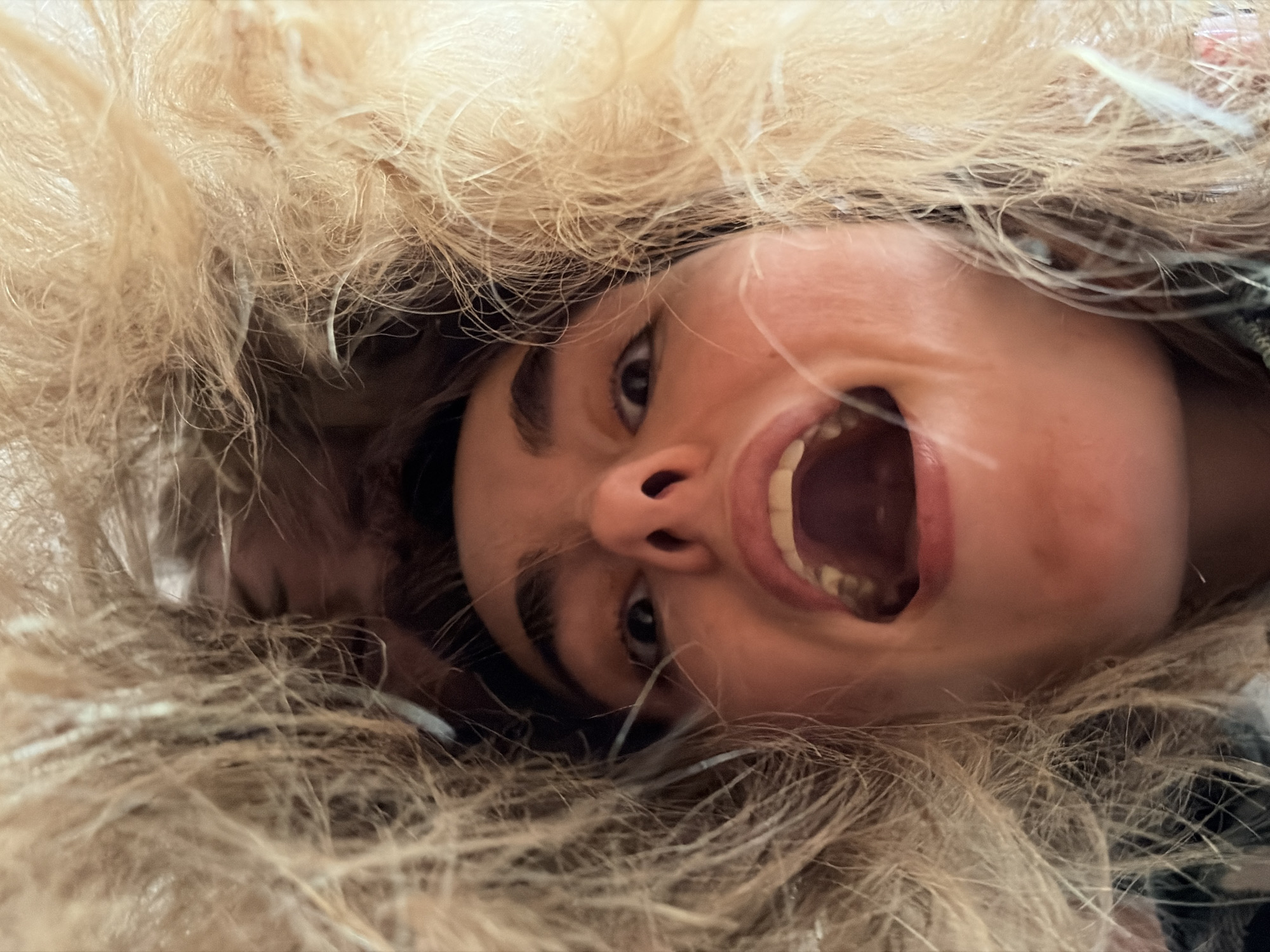 An upside-down close-up shot depicts a person wearing a hooded jacket. The hood's fur trim surrounds the person's face, obscuring parts of the background. The person's mouth is wide open, teeth visible. Their eyes are directed slightly upward. There's no indication of the location being Izhevsk, Russia.