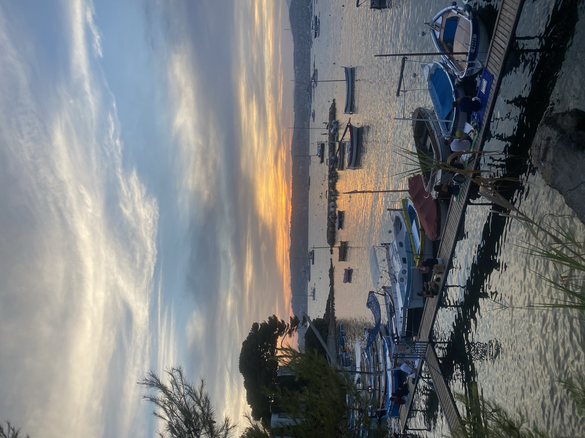 This image depicts a sunset scene at a harbor, likely in Antibes, France. Several small boats are docked along wooden piers, some of which have people sitting on them, enjoying the view. The sky is filled with wispy clouds, tinged with the warm colors of the setting sun, casting reflections on the calm water. A large pine tree and some foliage are visible in the foreground, adding a natural element to the otherwise man-made harbor. The distant shoreline is lined with hills and buildings, indicating an inhabited area. The overall atmosphere is peaceful and serene, with the soft light of dusk creating a tranquil ambiance.