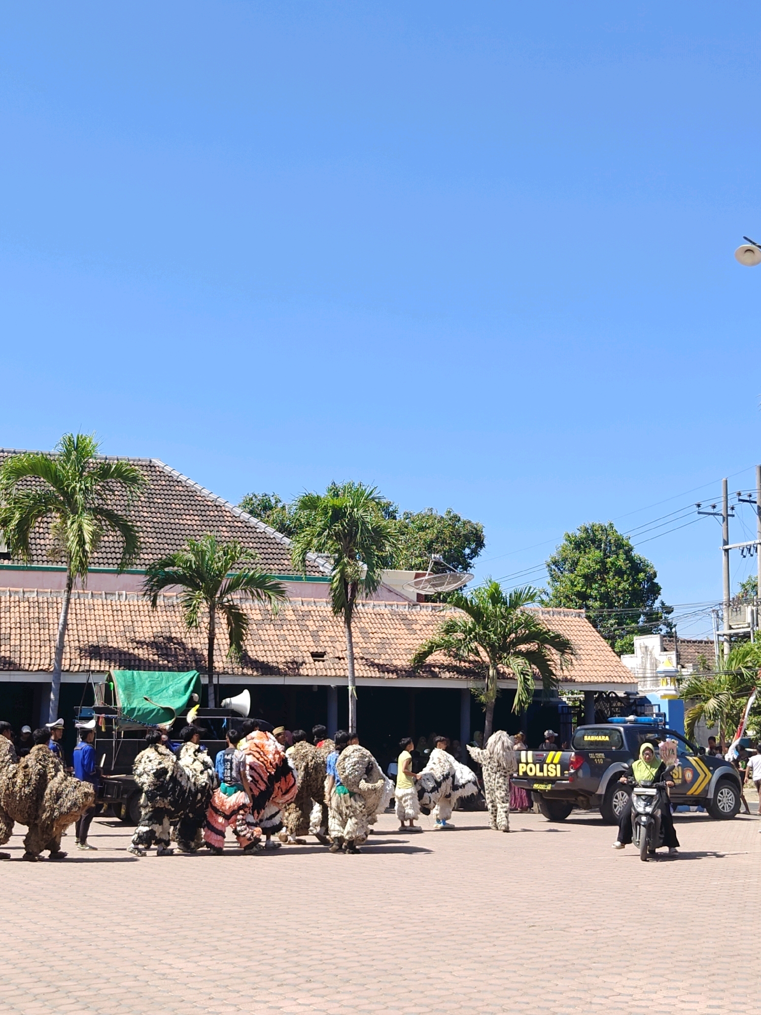 In Klampokan, Indonesia, a bright, clear sky forms the backdrop for a street scene featuring a procession of individuals in animal-like costumes. The ground is paved with brick. In the foreground, a group of people are walking, some in elaborate, fur-covered costumes. Behind them, a black police vehicle with "POLISI" visible on its side is parked. A person wearing a hijab is riding a motorcycle in front of the police vehicle. To the left, a stage or platform with a green tarp and a large speaker is visible, along with several palm trees and tiled-roof buildings. The overall impression is of a festive or cultural event taking place outdoors.