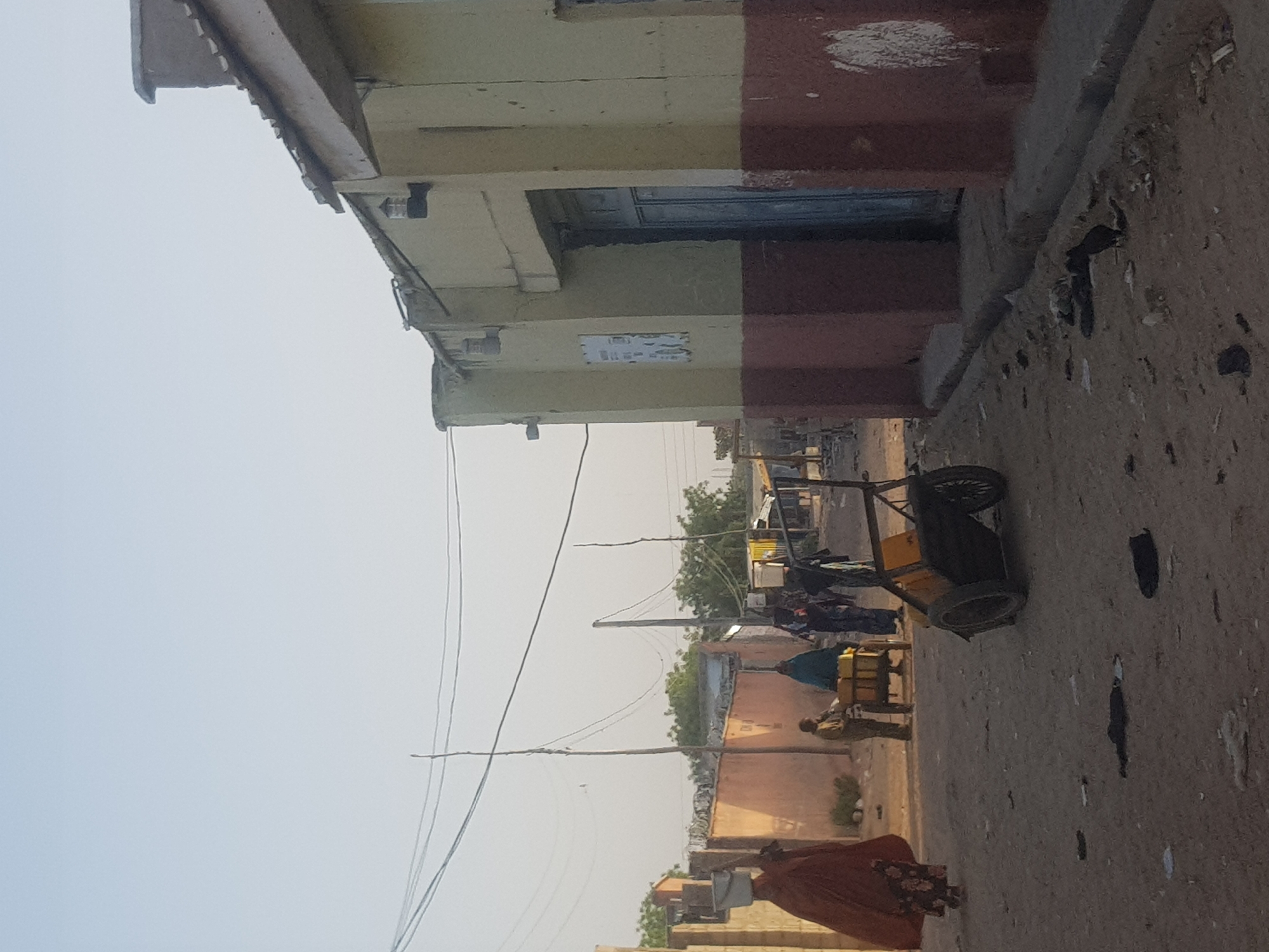 The image shows a street in Dabawa, Nigeria. The street is unpaved and has litter scattered along it. A small, yellow cart is parked on the side of the street. Several people are walking and standing nearby. Overhead power lines run along the street. Buildings of various heights line the street with painted walls and simple designs. The sky is overcast.