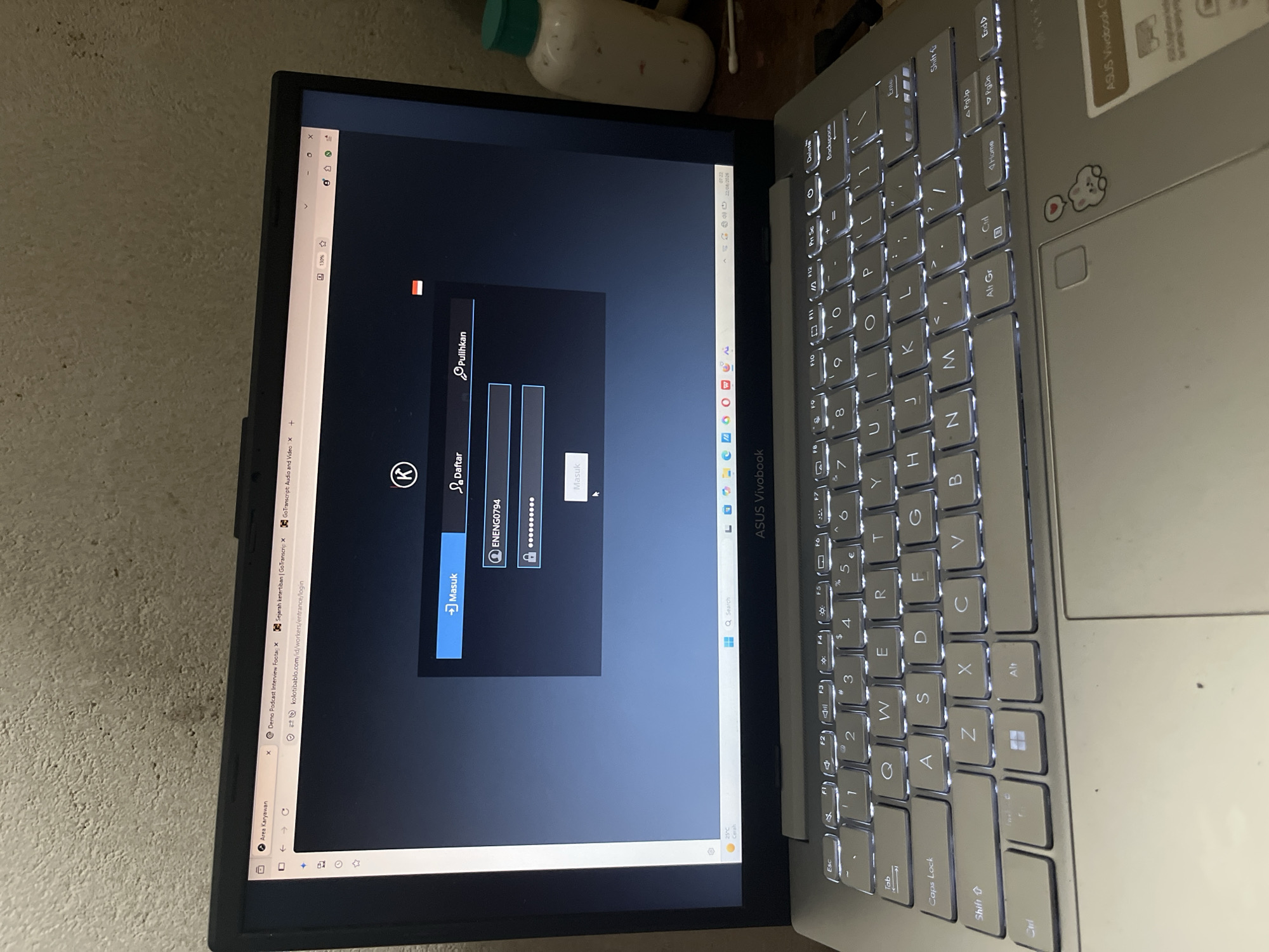 The image shows a laptop screen displaying a login page for a website. The website has a dark theme with the logo "K" prominently displayed at the top. There are fields for username and password, with "ENENG0794" entered in the username field and a series of dots representing the password. A button labeled "Masuk" (which means "Login" in Indonesian) is visible below the password field. To the right of the "Masuk" button, there are two options: "Daftar" (Register) and "Pulihkan" (Recover).

The laptop itself is an ASUS Vivobook, as indicated by the branding on the palm rest. The keyboard is illuminated. In the background, a wall with a textured surface is visible. To the right of the laptop, a white plastic bottle with a green cap and a cotton swab are present, suggesting a casual or domestic setting. The overall scene appears to be an indoor environment, likely a room, but the specific location within Meliau Hulu, Indonesia, cannot be determined from the image alone.