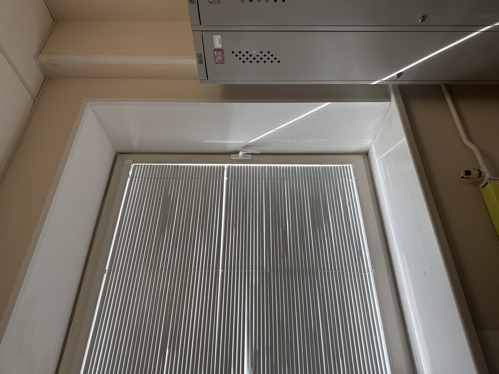 This media file captures an indoor scene, likely in Moscow, Russia, focusing on a window and overhead storage. No people are visible.
The primary subject is a window, fitted with vertical blinds that are partially drawn, allowing strong, diagonal shafts of natural sunlight to cut across the room. The window frame and handle are white. Above the window, a light-colored wall or ceiling meets a series of light grey, metallic cabinet or locker units mounted high on the wall. These units feature small square ventilation holes and two of them have locks.
Notable details include the bright sunlight, indicating a clear, sunny day, likely morning or late afternoon given the low angle of the light. On one of the grey cabinet units, a red-purple label with "36 A" is visible. Another cabinet unit has text in Cyrillic, "ПРАКТИКА", which translates to "Practice" or "Practika." To the right of the window, a wall features exposed white piping and a small yellow-and-black device, possibly a sensor or part of an alarm system, attached to the wall. The overall color palette is neutral, dominated by white, beige, and light grey. The environment appears functional, possibly an office, classroom, or technical area.