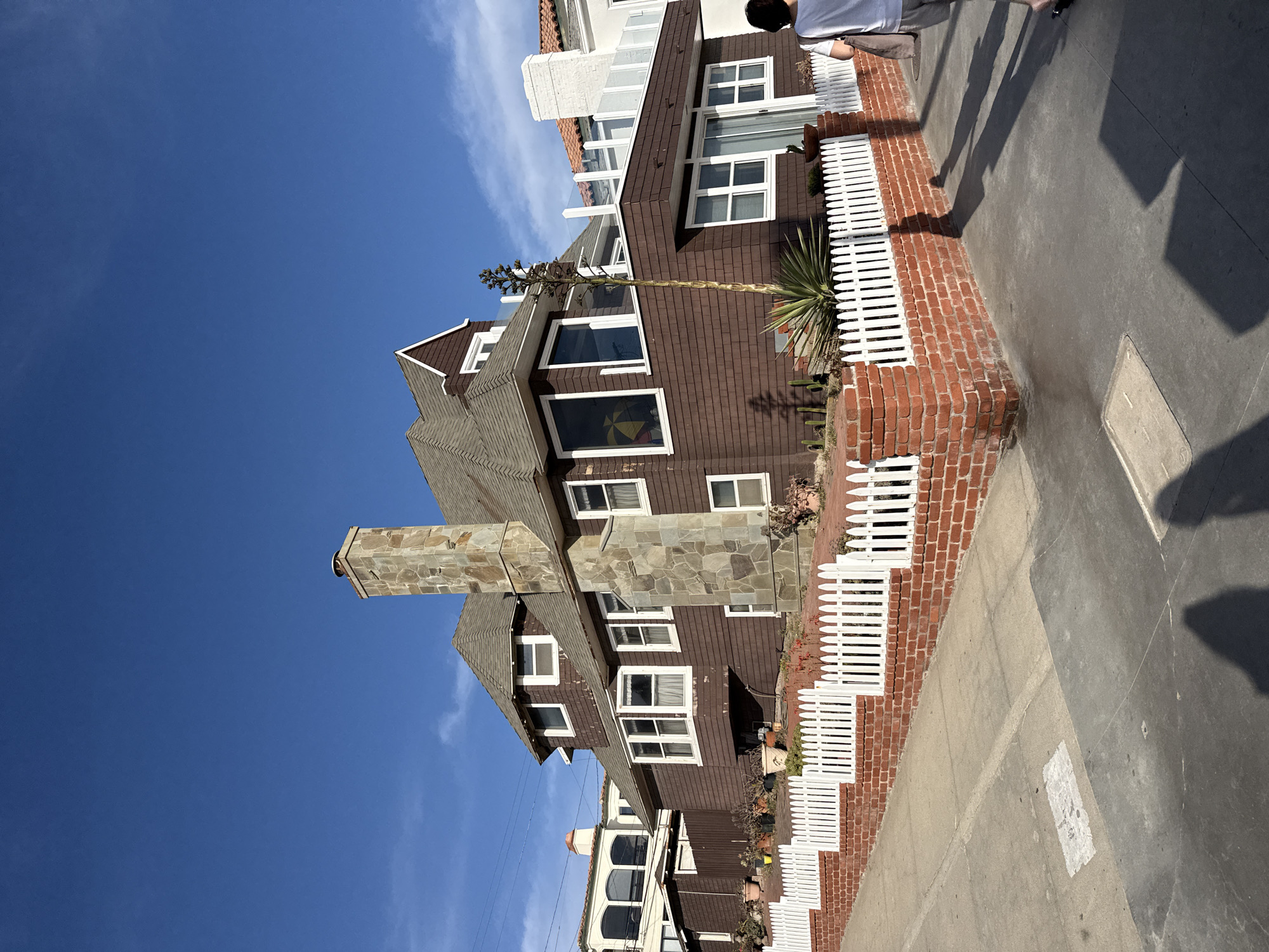 This image depicts a brown shingled house with a prominent stone chimney, located in Manhattan Beach, California, United States of America. The house features numerous windows with white trim and a steeply pitched roof. A white picket fence with a red brick base lines the front of the property. In front of the fence, various plants and succulents are visible, including a tall agave plant. To the left, other houses are partially visible. A person, viewed from behind, is walking on the sidewalk to the right of the frame, wearing a white t-shirt and grey shorts, carrying a tote bag. Shadows of people are cast on the sidewalk. The sky is clear and blue with a few wispy clouds.