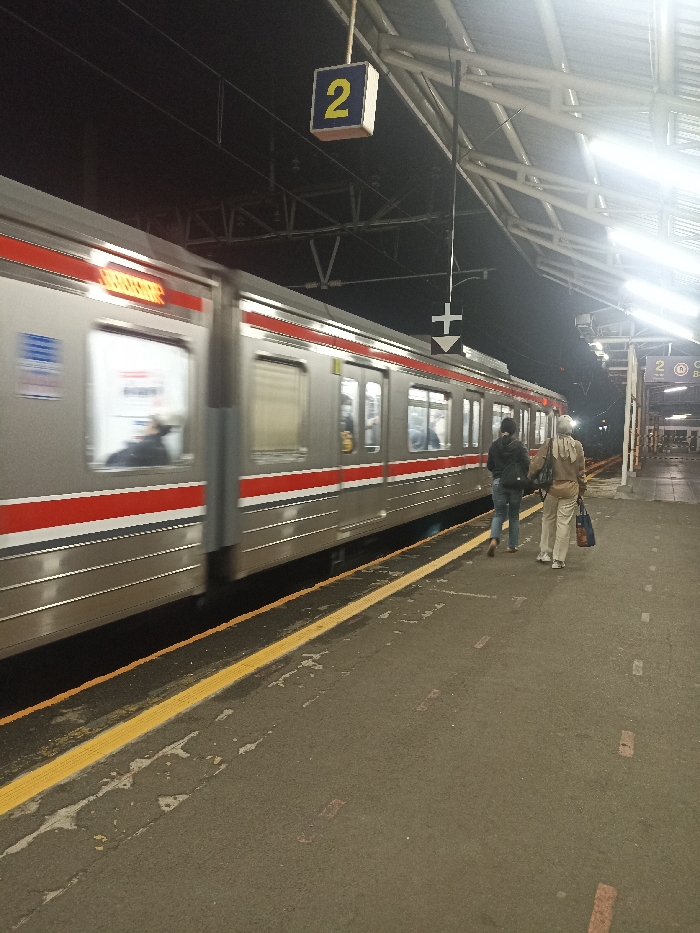 A train is moving away from the camera on the left side of the frame. There is a sign that says "2" above the train. The train is leaving the station. There are two people walking away from the train on the right side of the frame. The platform is made of concrete and there is a yellow line on the ground. The station is located in Bojong Gede, Indonesia at the coordinates -6.494 latitude and 106.795 longitude. There is a sign with a downwards pointing arrow. The environment is nighttime. The lighting is dark and there are streetlights.