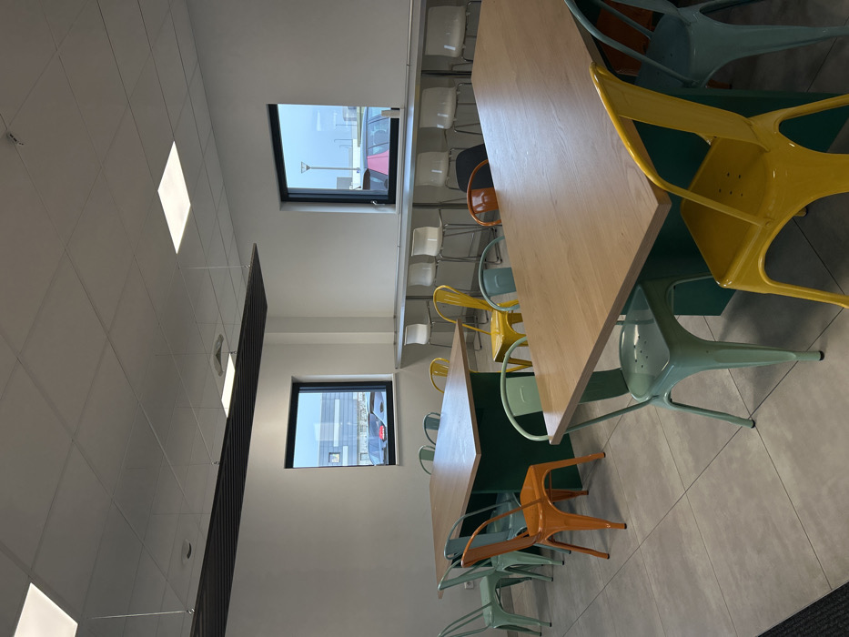 The image displays an unoccupied modern interior space, identified as being located in Reichstett, France. The room features white walls, a light grey tiled floor, and a suspended acoustic tile ceiling containing multiple square LED lighting panels. A long, rectangular black slatted acoustic panel is suspended from the ceiling in the foreground-left.
Two large, rectangular tables with light wood-colored tops and dark green bases are positioned in the center and foreground. These tables are surrounded by numerous metal chairs in bright colors, including mint green, orange, and yellow.
Along the far wall, a long, narrow counter-height bar with a light grey surface extends beneath two rectangular windows, each framed in black. Several white bar stools with silver bases are arranged along this counter.
Through the windows, an exterior view reveals an industrial or commercial setting, including visible buildings and parked vehicles, under an overcast sky. No individuals are present within the depicted scene.
