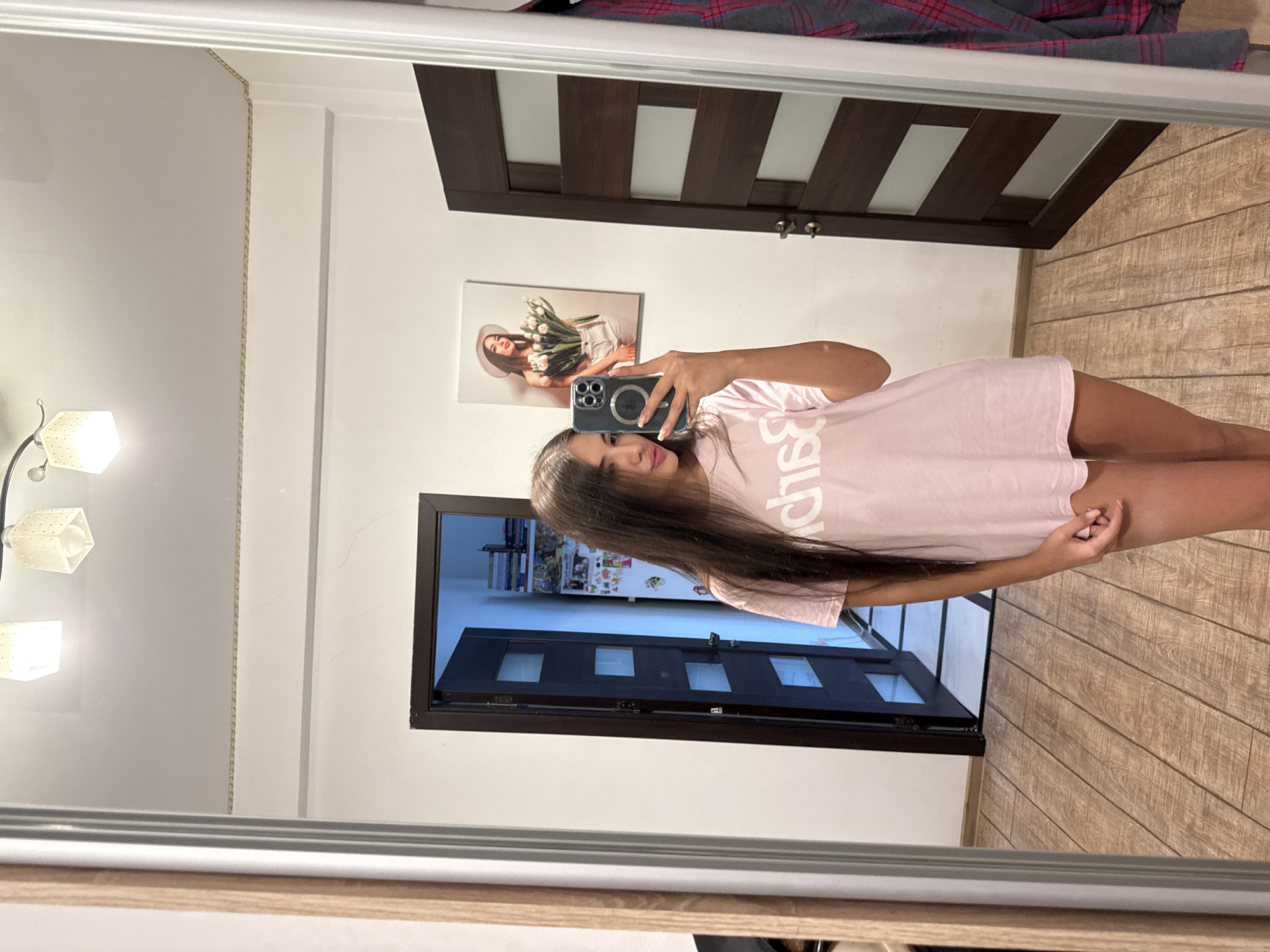 A young woman with long dark hair takes a selfie in a mirror. She is wearing a pale pink t-shirt and is standing on a wooden floor. Behind her, there are two dark wood doors, one slightly ajar revealing a glimpse of shelves and books. A framed picture of a woman holding tulips hangs on the wall. The scene appears to be indoors, possibly in a home or a hotel room in Chișinău, Moldova.