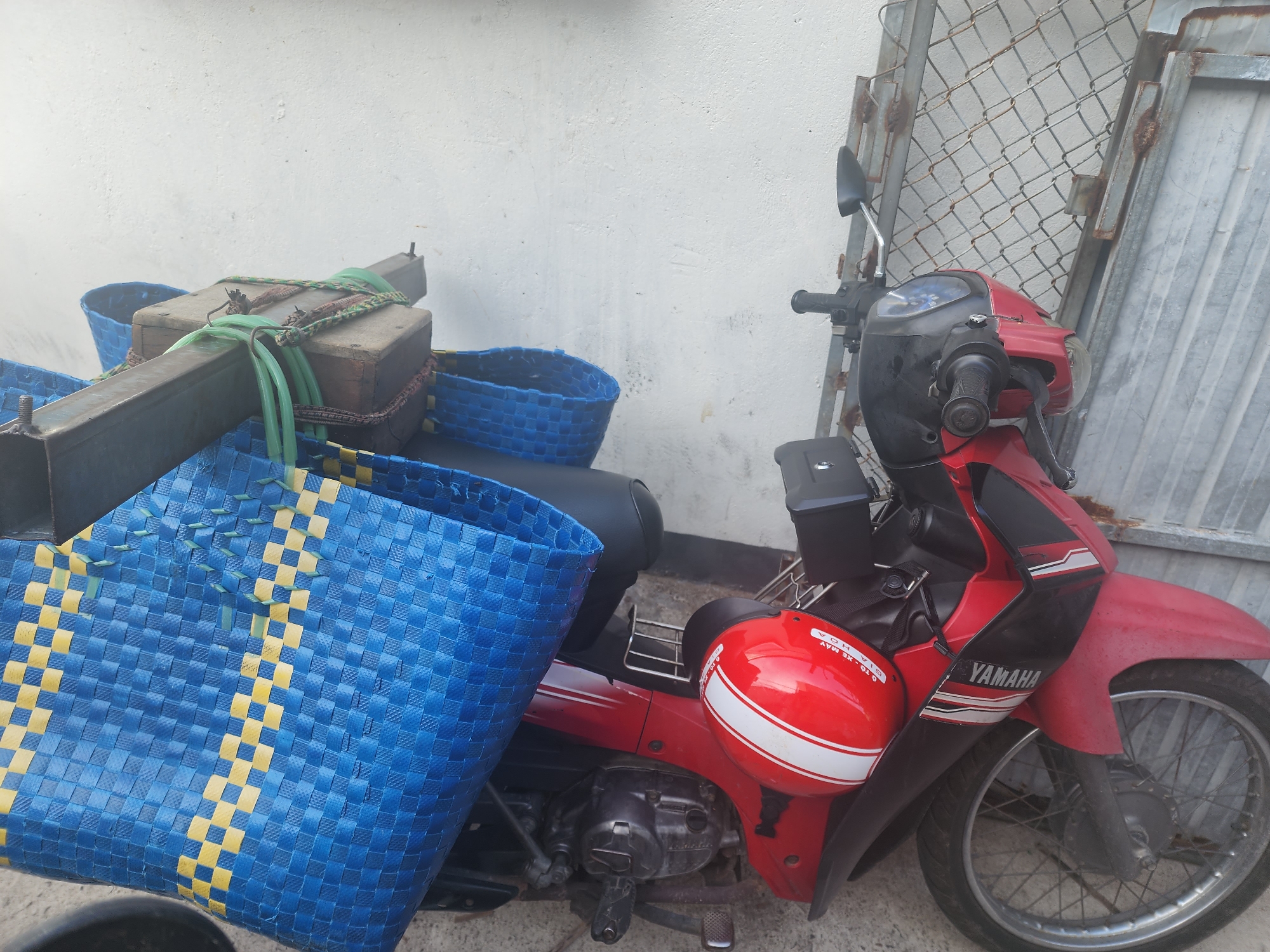 The image shows a red Yamaha motorcycle parked next to a white textured wall, likely in an outdoor urban setting such as Phu Thuy Ward, Vietnam. A large blue and yellow woven basket is attached to the rear of the motorcycle, and another smaller blue basket is visible behind it. A black seat and part of the motorcycle's engine are visible. A red helmet with white stripes is placed on the front of the motorcycle, near the handlebars, which are black. A metal fence, partially rusted, is situated behind the motorcycle and the wall. The ground appears to be concrete or pavement. There are no people in the frame. The lighting suggests it is daytime.