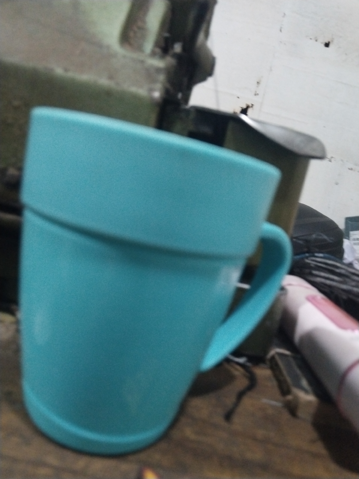 A close-up, blurry, outdoor, medium shot captures a turquoise mug in the foreground. The mug is positioned on a wooden surface, likely a table. To the right of the mug, a partially visible pink and white object, possibly a blender, rests on the surface. In the background, a portion of a large, green, industrial-looking machine is visible, along with a dark-colored, cylindrical object. The overall environment appears to be somewhat cluttered and dimly lit, suggesting a workshop or storage area. The image is taken from a slightly elevated angle. There are no people or discernible human interactions present in the scene. The location is not specified beyond "Unknown, Indonesia."