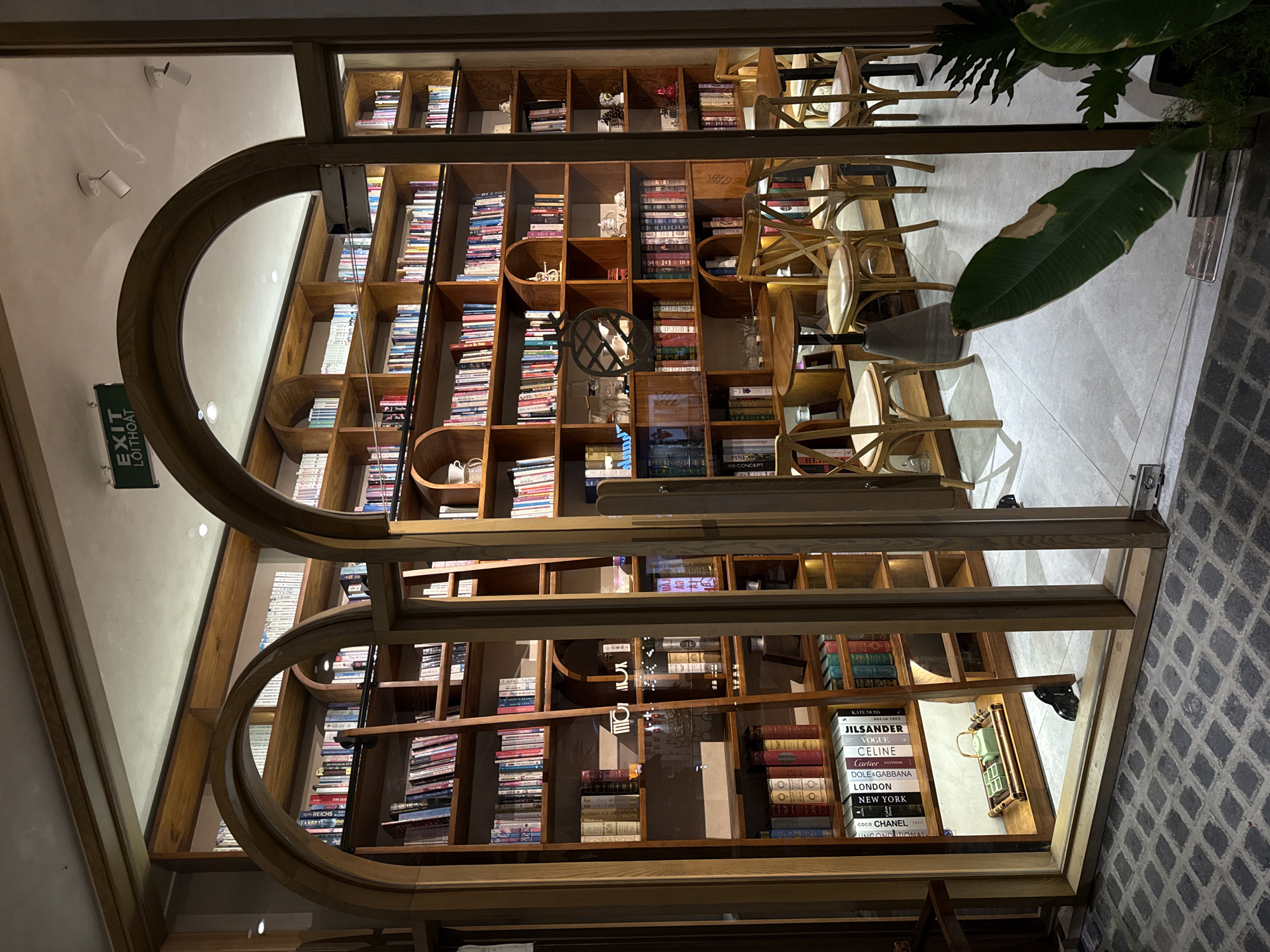 This image shows a wide shot of a bookstore interior, captured from a low angle looking upwards, and the image is rotated 90 degrees counter-clockwise. The primary subject is a large, multi-tiered wooden bookshelf filled with books. The shelves are organized into numerous cubby-like compartments, with books neatly stacked within. The books themselves vary in size and color, creating a visually rich and textured display. Interspersed with the books are some decorative items and what appear to be empty shelves.
In the foreground, a large, curved wooden archway frames part of the bookshelf, suggesting a unique architectural design for the space. Outside the bookshelf, chairs are visible, some stacked and others arranged, indicating a seating area for patrons. The floor is tiled with a patterned mosaic. A lush green plant with a large leaf is positioned in the upper right portion of the frame, adding a touch of nature to the indoor setting.
A prominent green sign with white text reads "EXIT LỐI THOÁT," indicating an exit route in Vietnamese. Various book titles are legible on the spines of some books, including "JIL SANDER," "VOGUE," "CELINE," "Cartier," "DOLE&GABBANA," "LONDON," "NEW YORK," "COCO CHANEL," and "PARIS." The lighting suggests an indoor environment, possibly during the day given the brightness, but without distinct shadows or sunlight. The overall impression is that of a well-stocked and inviting bookstore, with a focus on the visual appeal of the books and the architectural elements of the space.