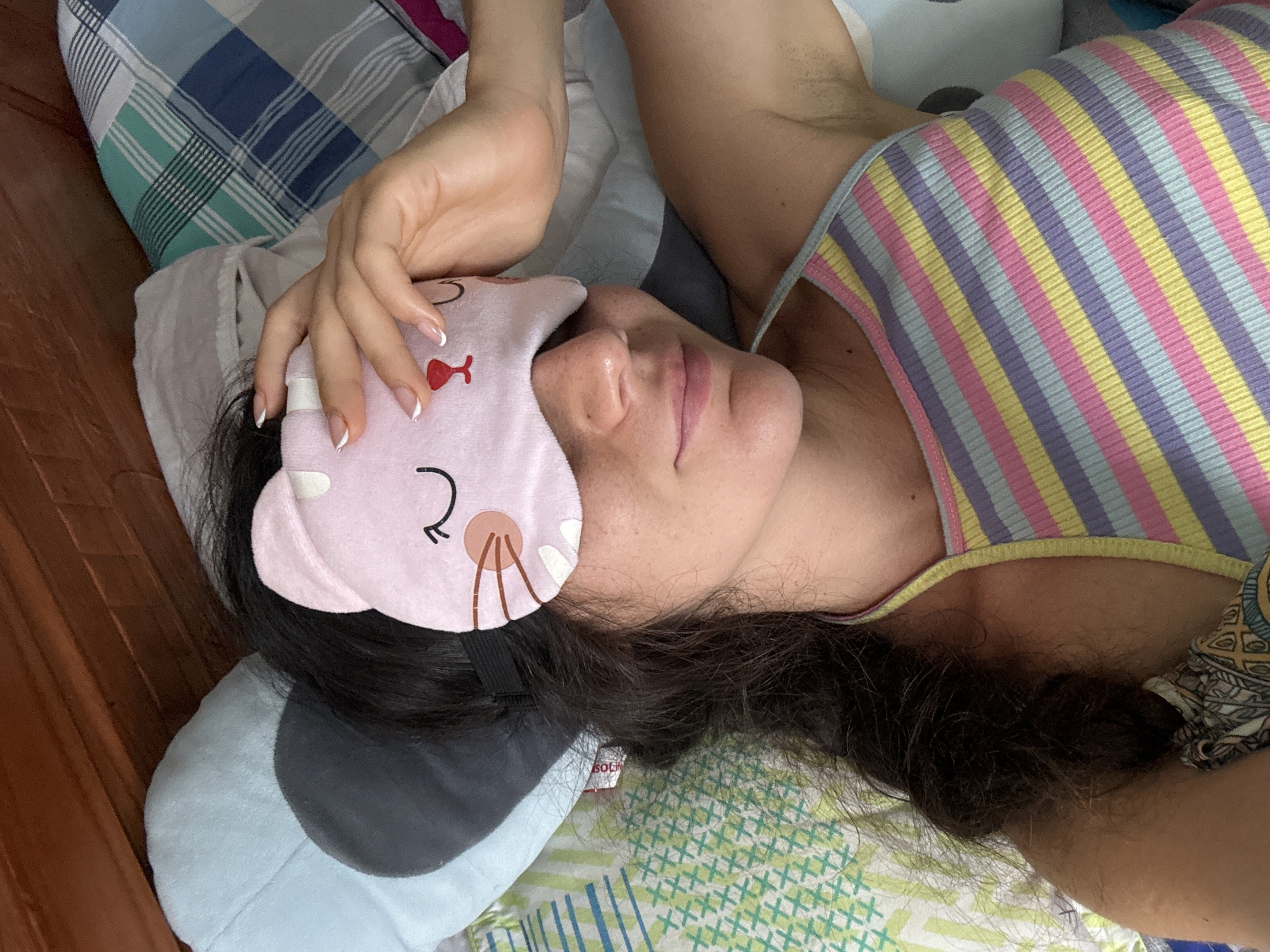 A person is lying in bed with a pink cat-shaped eye mask covering their eyes. Their left hand is resting on top of the eye mask. They are wearing a multicolored striped tank top. The background shows pillows and a wooden headboard, consistent with a bedroom environment. The scene is indoors.