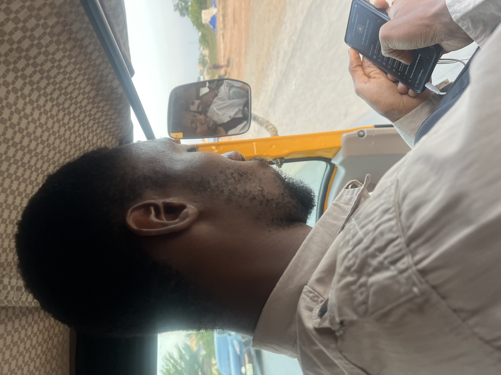 The photograph shows a man inside a vehicle, possibly a taxi or auto-rickshaw. He is dark-skinned, with short black hair and a beard. He is wearing a light-colored, button-down shirt. He is holding a black smartphone in his hands, connected to a white charging cable. The phone's screen is visible, displaying a profile and possibly a menu. A side mirror is visible in the background, reflecting the man's face and surroundings, which include other people and objects that are unclear due to the mirror's distortion. Through the vehicle's window, outside scenes are visible, with other vehicles. Based on the prompt, the location is identified as Karum, Nigeria.