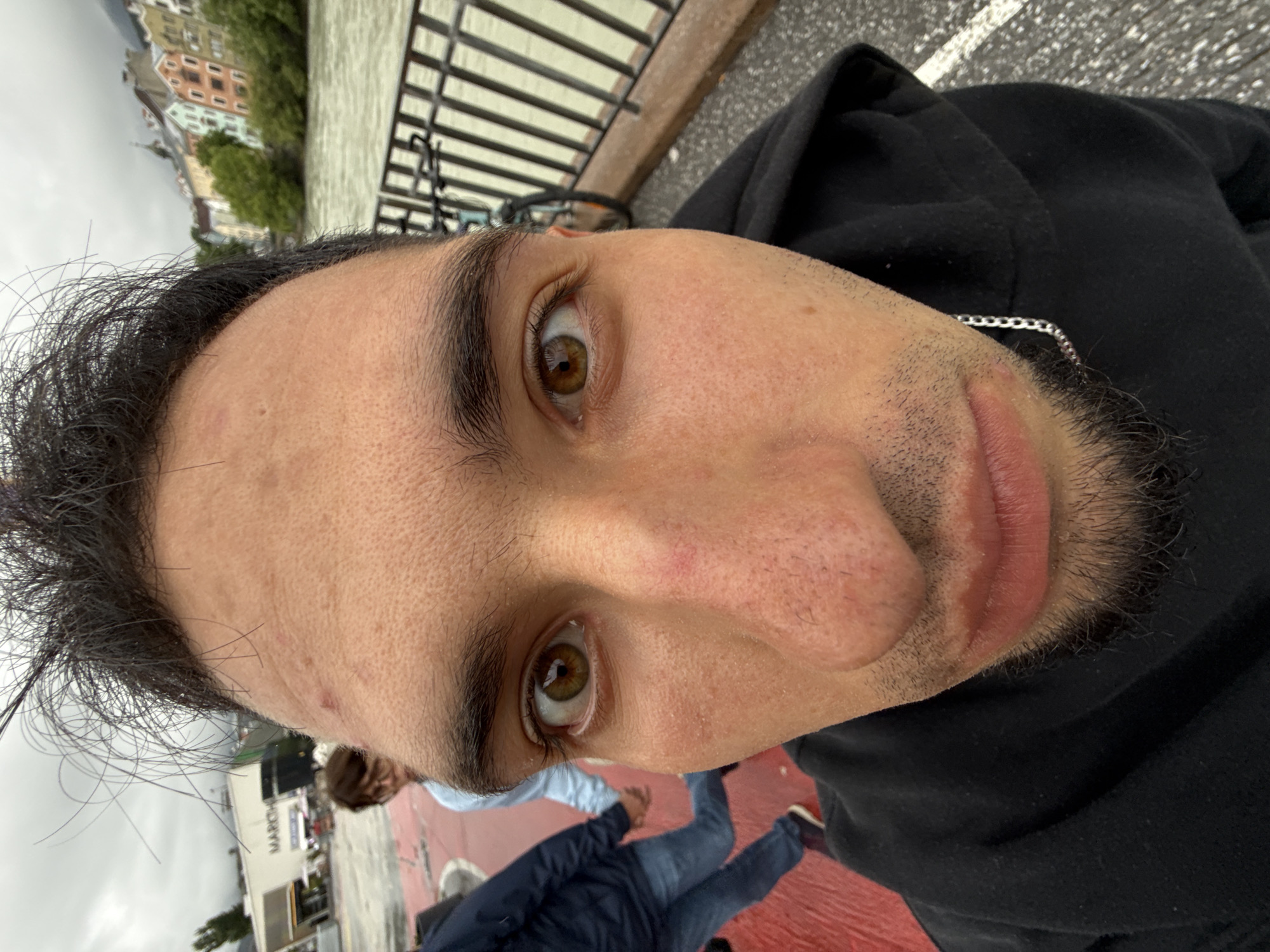 The image is a close-up, fisheye-lens selfie taken outdoors in Innsbruck, Austria. The foreground is dominated by the face of a young man wearing a black hoodie. His features are distorted by the wide-angle lens, with his forehead and nose appearing significantly enlarged. He has dark, unkempt hair, brown eyes, and a light beard. A silver chain is visible around his neck.
In the background, a river flows alongside a paved walkway with a black metal railing. Across the river, a row of colorful, historic buildings is visible on a hillside, characteristic of the city of Innsbruck. To the left of the frame, two other individuals are partially visible. One, wearing a light blue jacket, is holding hands with another person dressed in dark clothing and blue jeans. Further in the background, a building with a sign that reads "MARCH" can be seen. The sky is overcast, suggesting a cloudy day. The overall scene depicts a casual moment captured on a bridge or walkway overlooking the Inn River in Innsbruck.