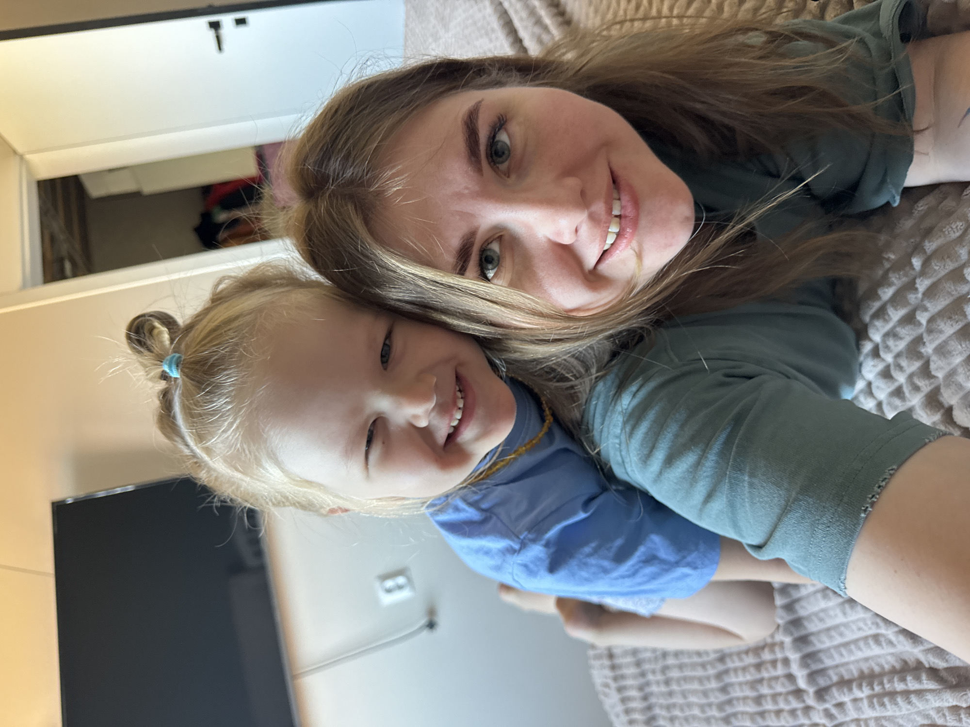 A woman and a child are lying on a bed, smiling at the camera. The woman has long, light brown hair and is wearing a green shirt. The child has blonde hair pulled back into a ponytail with a blue hair tie and is wearing a blue shirt and an amber necklace. A television and an electrical outlet are visible in the background. The location is inside a building in Ферма, Russia.