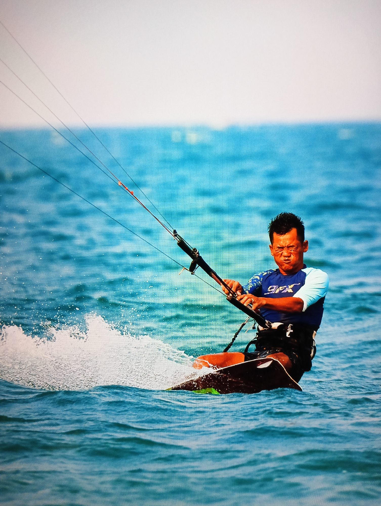 A medium shot depicts a man kitesurfing in the ocean. The man is in the center, wearing a blue and white long-sleeved shirt and black shorts, holding onto a black kite bar with both hands. His face shows signs of exertion. He is riding a kiteboard with a green edge and a dark top. Spray is visible to his left, emanating from the kiteboard hitting the water. Thin black lines extend from the kite bar upward and out of frame. The water is a clear turquoise color, and the background consists of a blurry horizon of blue ocean meeting a light sky. The location is Nha Trang Ward, Vietnam.