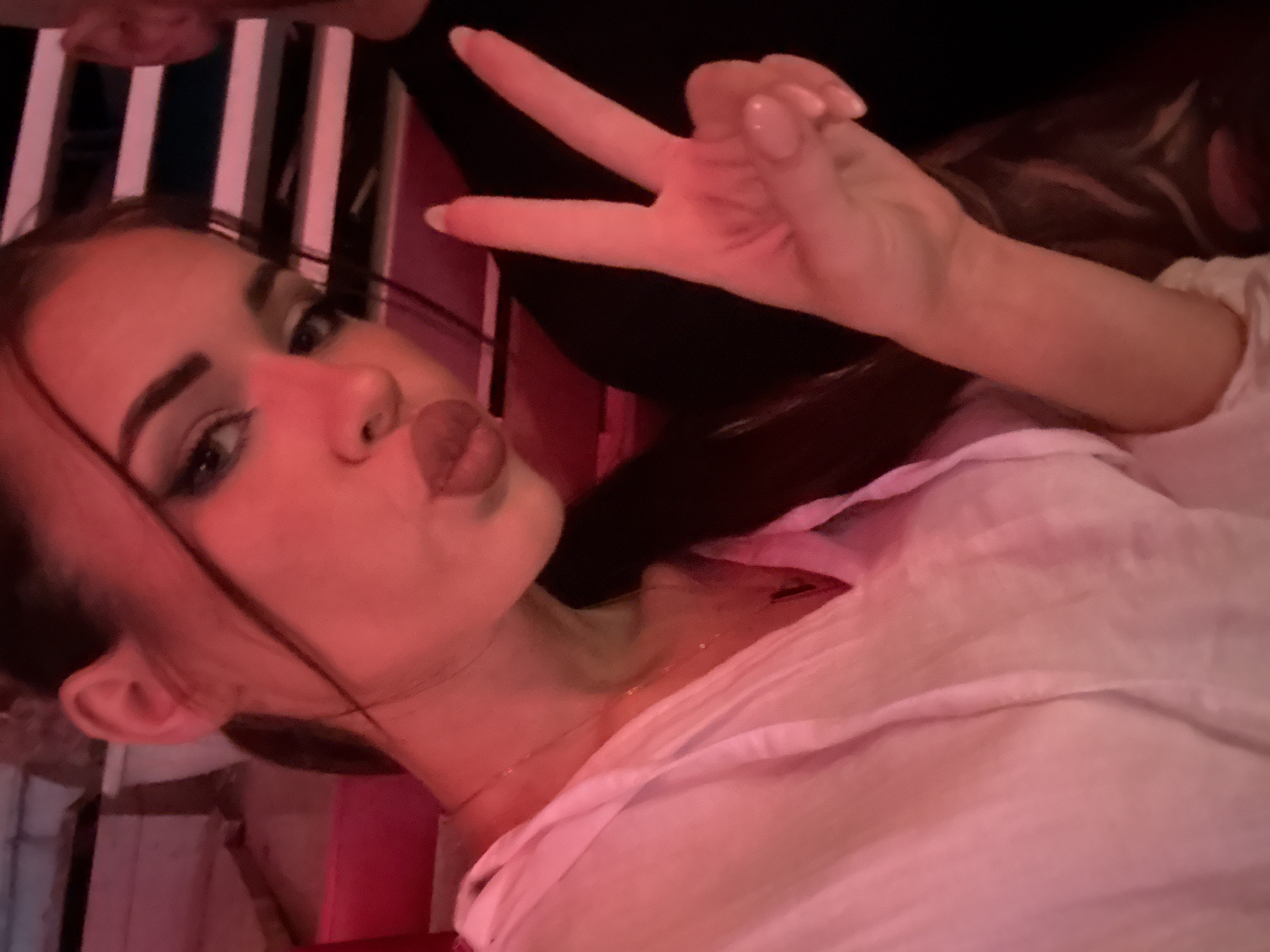 A portrait-oriented photograph captures a young woman posing in a dimly lit indoor setting in Plovdiv, Bulgaria, likely at night. She is positioned diagonally across the frame, with her head tilted, and her expression features dark eyeliner and puckered lips in a "duck face." Her dark hair is visible, and she is wearing a light-colored, seemingly white, casual top with a thin necklace. Her right hand is raised, making a peace sign with her manicured index and middle fingers.
The scene is bathed in a strong red and pink artificial light, casting a warm, atmospheric glow. The background is somewhat blurred but shows vertical stripes of dark and light colors, possibly part of the venue's decor. A small portion of what appears to be another person's forehead is visible at the very top edge of the image. The woman's playful pose and confident expression suggest she is enjoying a social event or outing. No visible text is present.