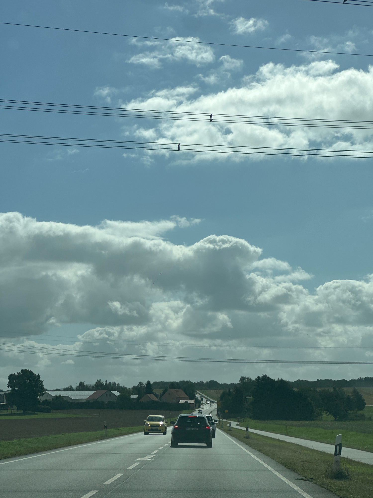 An eye-level shot captures a highway scene near Radibor - Radwor, Germany, under a sky filled with dramatic cumulus clouds. The highway stretches forward, marked with white dashed lines, with a paved shoulder on the right. Several vehicles are visible on the road, including a black SUV in the foreground, a yellow compact car further ahead, and other cars in the distance, suggesting moderate traffic. On the left side of the highway, a field transitions into a rural landscape with houses and trees, indicating a suburban or rural area. Overhead, power lines crisscross the frame, supported by poles and insulators, adding a man-made element to the natural scenery. The overall impression is a typical day on a German highway, blending infrastructure with the surrounding countryside.