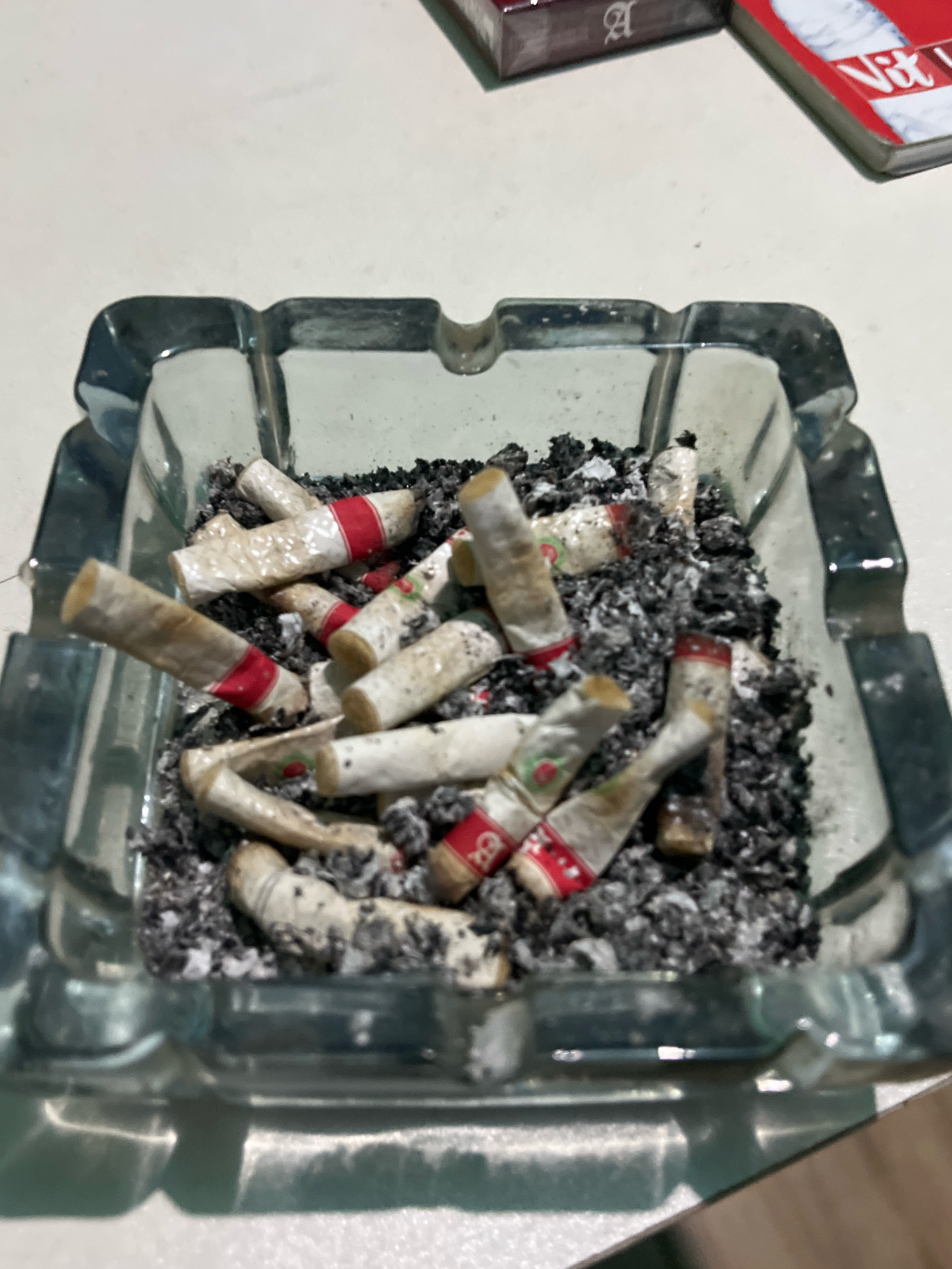 The image shows a close-up, overhead view of a glass ashtray filled with cigarette butts and ash. The ashtray is square with rounded corners and appears to be made of clear glass, with a textured pattern on its exterior. Inside, there are numerous partially smoked cigarettes, many with red and white bands. The ash is dark gray and scattered amongst the butts. In the background, out of focus, are two cigarette packs. One is dark colored with a white stylized "A" logo, and the other is red and white with text that includes "Vit". The scene is set on a light-colored, flat surface, likely a table. There is no indication of the city of Talang Kelapa, Indonesia within the image itself.
