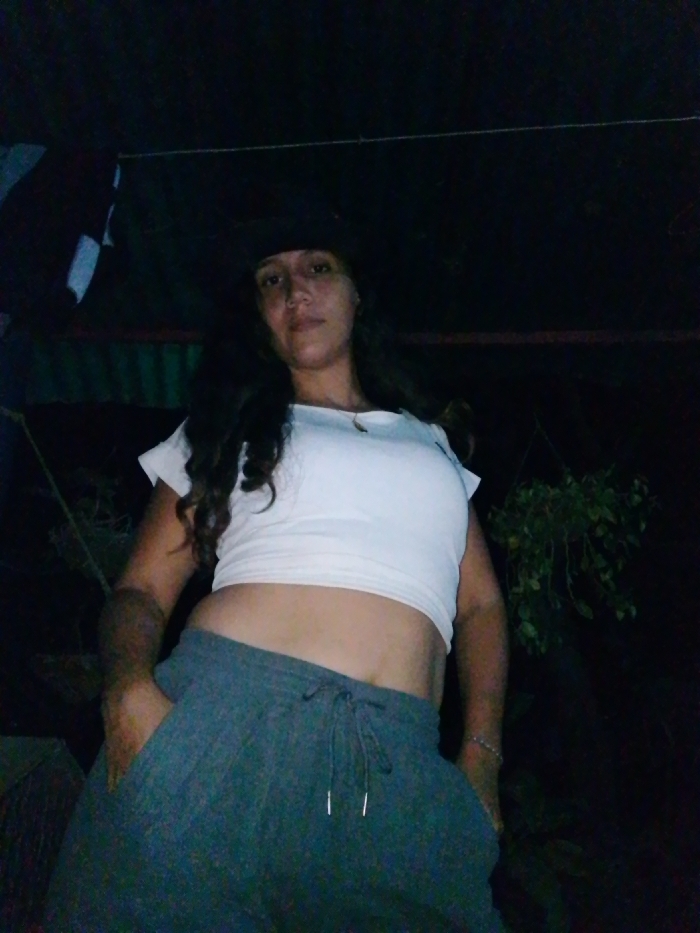 The image displays a single female subject, captured from a low-angle perspective in a dimly lit environment. The subject, centrally positioned, has long, dark, wavy hair and is wearing a dark cap, a white short-sleeved crop top that exposes her midriff, and dark grey sweatpants with a visible drawstring. Her right hand (viewer's left) is placed in her pocket, and a light-colored bracelet is visible on her left wrist (viewer's right). She looks directly at the camera with a neutral facial expression. The background features various dark elements: a vertical striped pattern is visible to the left, a thin horizontal line (possibly a wire or string) spans across the upper middle, and dark foliage or plant material is present on the right side. The ambient light is low. The specific location within Juan Griego, Venezuela, is not discernible from the visual evidence in the image.