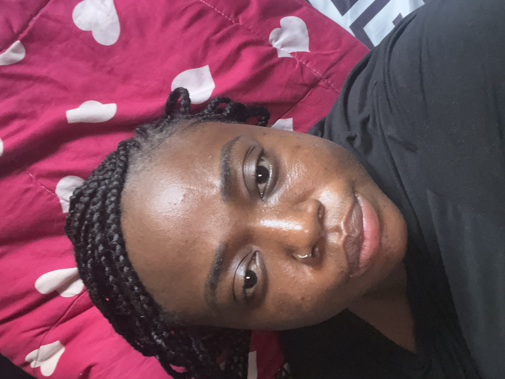 The image displays a close-up, eye-level selfie of an individual, likely a young adult or adult female, lying down indoors. The person has dark skin, braided hair, and is wearing a black top. A small silver hoop nose ring is visible in their left nostril. Their expression is neutral to relaxed, and they are looking directly at the camera. Their skin appears to have a dewy or reflective finish.
The background primarily consists of bedding. A vibrant pink comforter or duvet patterned with white hearts covers a significant portion of the frame around the person. In the upper right corner, a small section of another piece of bedding, possibly a pillow or sheet, with a black and white geometric pattern is visible. The scene suggests a bedroom setting.
The activity taking place is a self-portrait (selfie). Time of day and weather are not discernible from the indoor setting. There is no visible text in the image. The provided context indicates the location is Abuja, Nigeria.