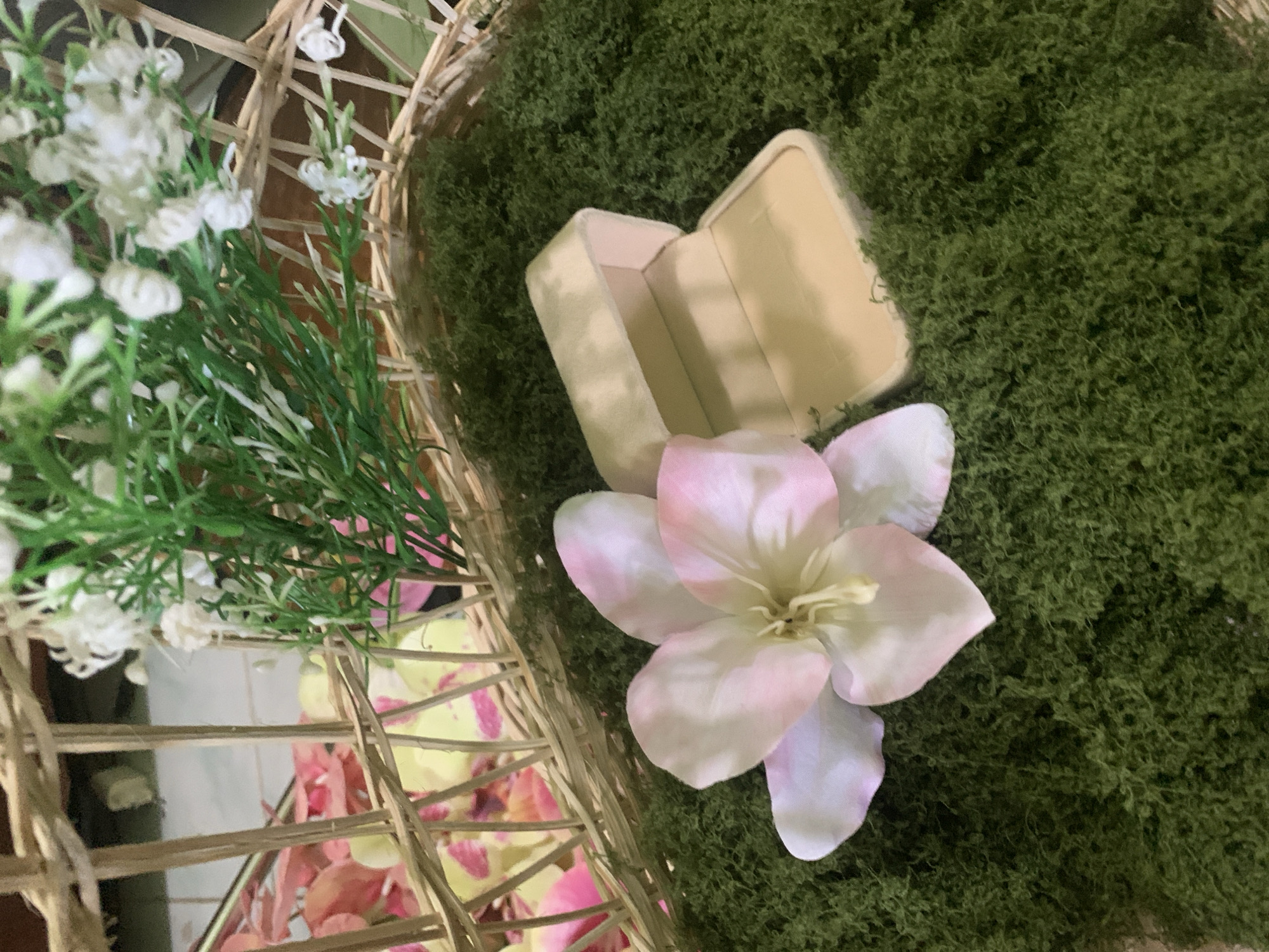 In a woven basket, a pale pink and white flower rests on a bed of green moss. Beside the flower, an open, light beige ring box sits on the moss. In the background, more flowers and foliage are visible, some in pink and yellow, and some green with small white blossoms. The scene appears to be indoors. The provided context does not contain information about the specific location of Kuala Pembuang, Indonesia, or any details about people, actions, or interactions.