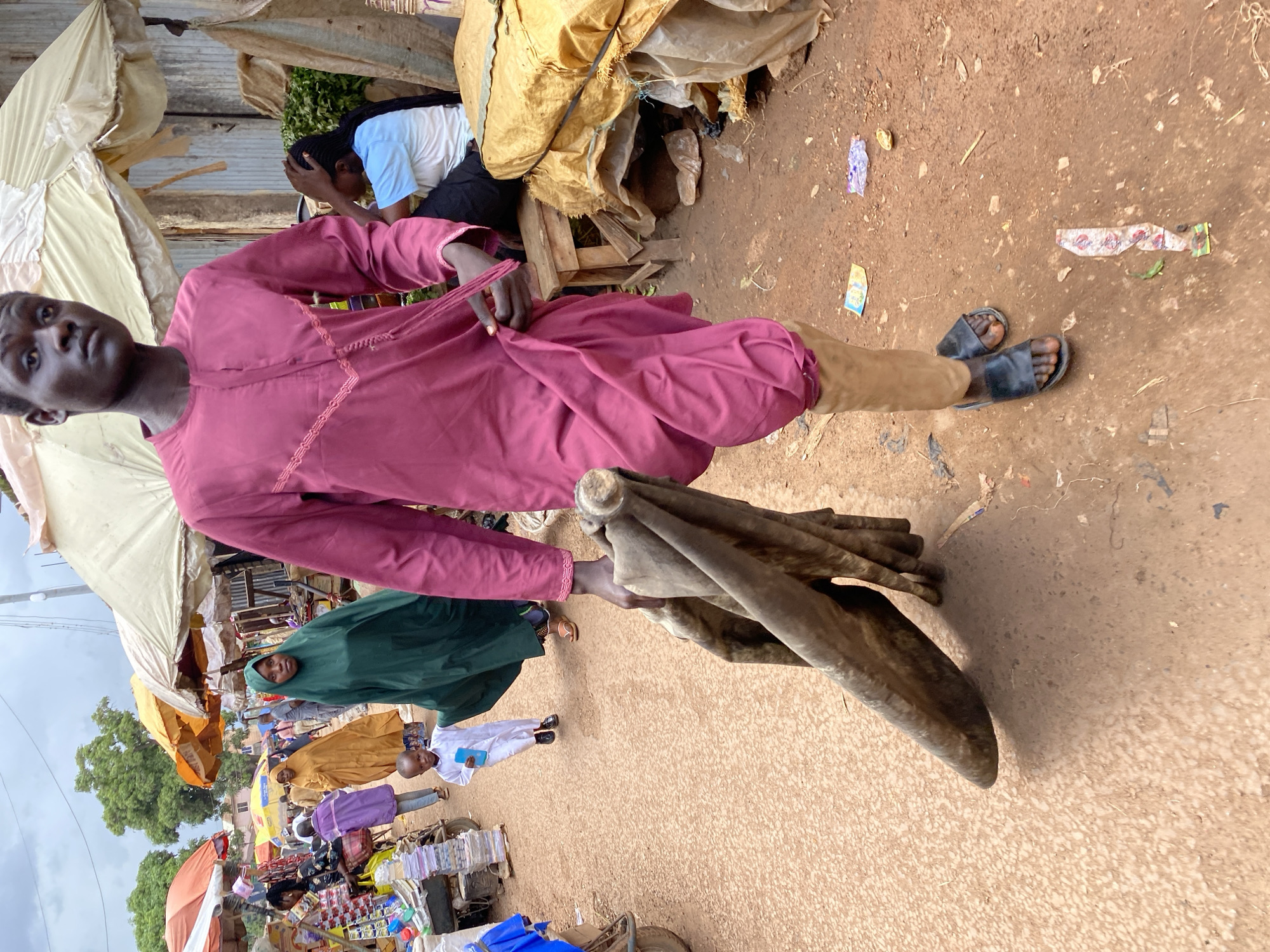 In Bukuru, Nigeria, a young man in a maroon tunic and tan leg coverings walks forward carrying a large, rolled-up piece of dark brown material. He is in the foreground, facing the viewer. Behind him, a dirt path is visible, lined with market stalls and people. To his left, a woman in a green hijab and another in a yellow garment are walking away. Further back, a young child in a white outfit is also walking. The market appears to be outdoors, with various tents and awnings providing shade. The ground is made of reddish-brown dirt, and there are some scattered pieces of trash. The overall scene depicts a busy marketplace.