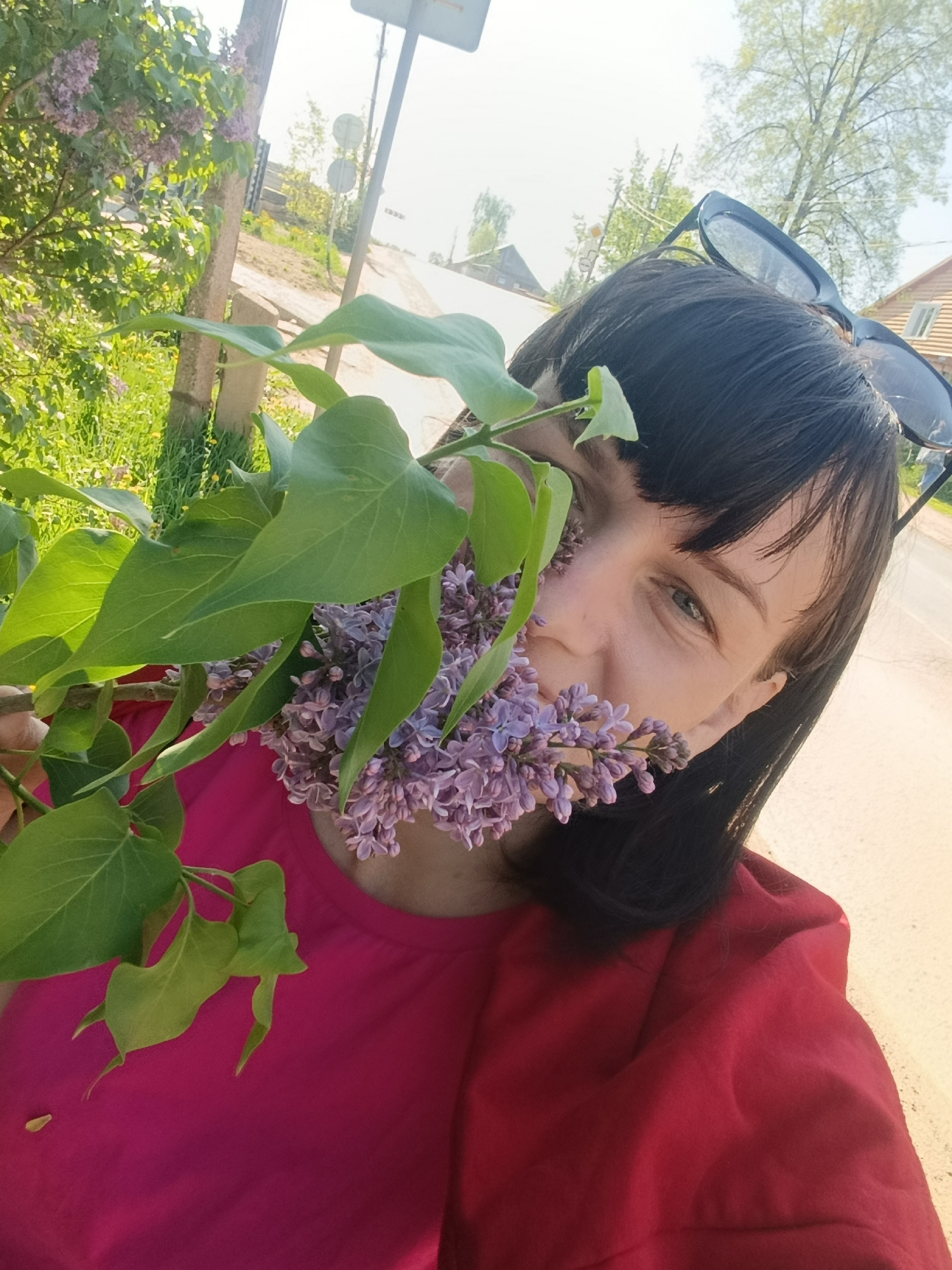A medium-shot captures a woman holding a branch of lilacs, partially obscuring her face. Her dark hair is styled with bangs, and she wears sunglasses perched on her head. She is dressed in a red top. The setting appears to be outdoors, near a road with sparse vegetation. In the background, there are houses and trees, indicative of a rural or suburban environment. The photograph was taken in the city of Фоки, Russia.