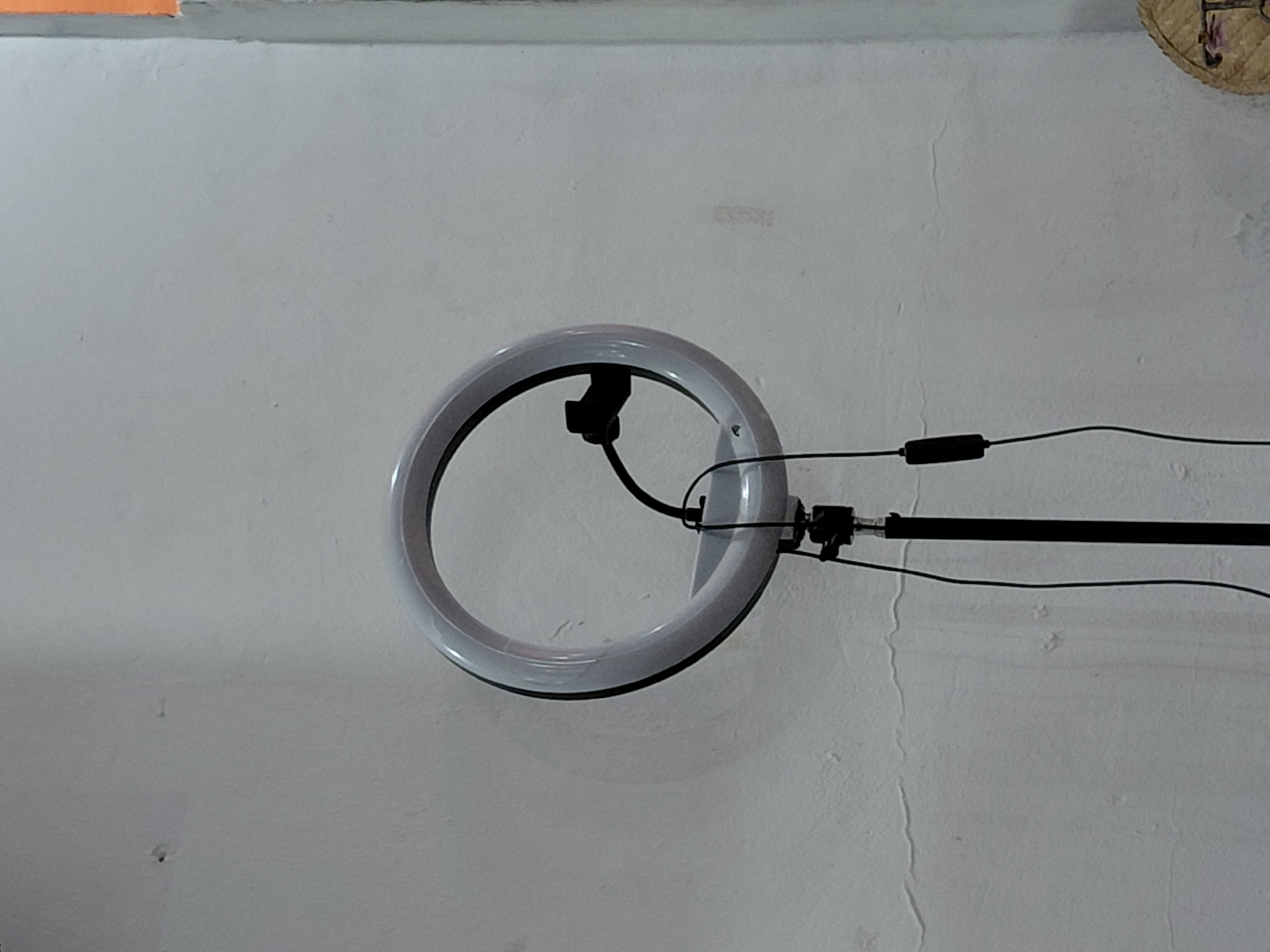 The image displays a white ring light illuminated and mounted on a black stand, set against an off-white wall. The ring light, circular in shape with a black interior, is the central focus, emitting a soft glow. A black cable extends from the light, connecting to a small rectangular control unit situated on the black stand, which stretches horizontally to the right side of the frame.
The wall behind the equipment exhibits a textured surface with minor imperfections and faint marks, suggesting wear or an unpolished finish. In the top left corner, a narrow segment of an orange-colored surface is visible, possibly a ledge or a corner of an adjacent structure. The upper right corner reveals a small portion of a woven, possibly straw or wicker, object, suggesting a decorative element or household item.
The lighting in the photograph appears to be primarily sourced from the ring light itself, highlighting the equipment. No individuals are present, and no specific activity is taking place; the image is a static portrayal of a lighting setup. There is no discernible text within the image. The scene is located in Santa Ana, Venezuela.